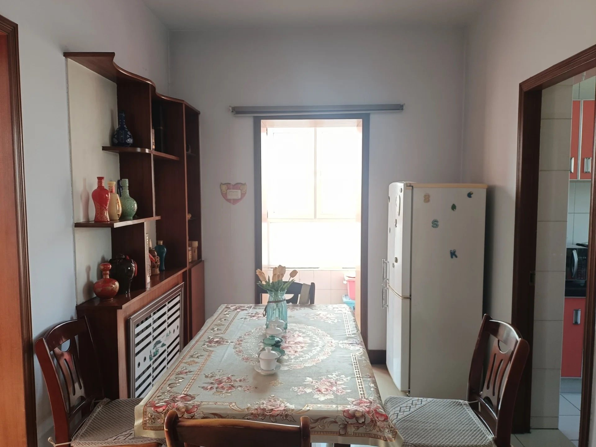 温馨小厨房，白墙配窗，中央餐桌四椅环绕，点缀花瓶装饰。厨房内有餐具、两杯、碗和一瓶，冰箱位于角落。自然光透过窗户，营造明亮舒适的用餐或聚会氛围。关键字:小厨房 白墙 窗户 餐桌 椅子 花瓶 餐具 杯子 碗 瓶子 冰箱 自然光 用餐氛围