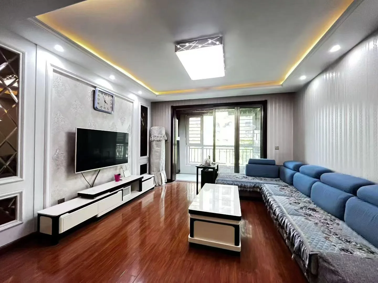 宽敞的客厅，硬木地板，左侧有大屏幕电视，右角摆放沙发，中央有咖啡桌，两扇窗户引入自然光，窗边各有一把椅子。关键字:宽敞客厅 硬木地板 大屏幕电视 沙发 咖啡桌 自然光 椅子