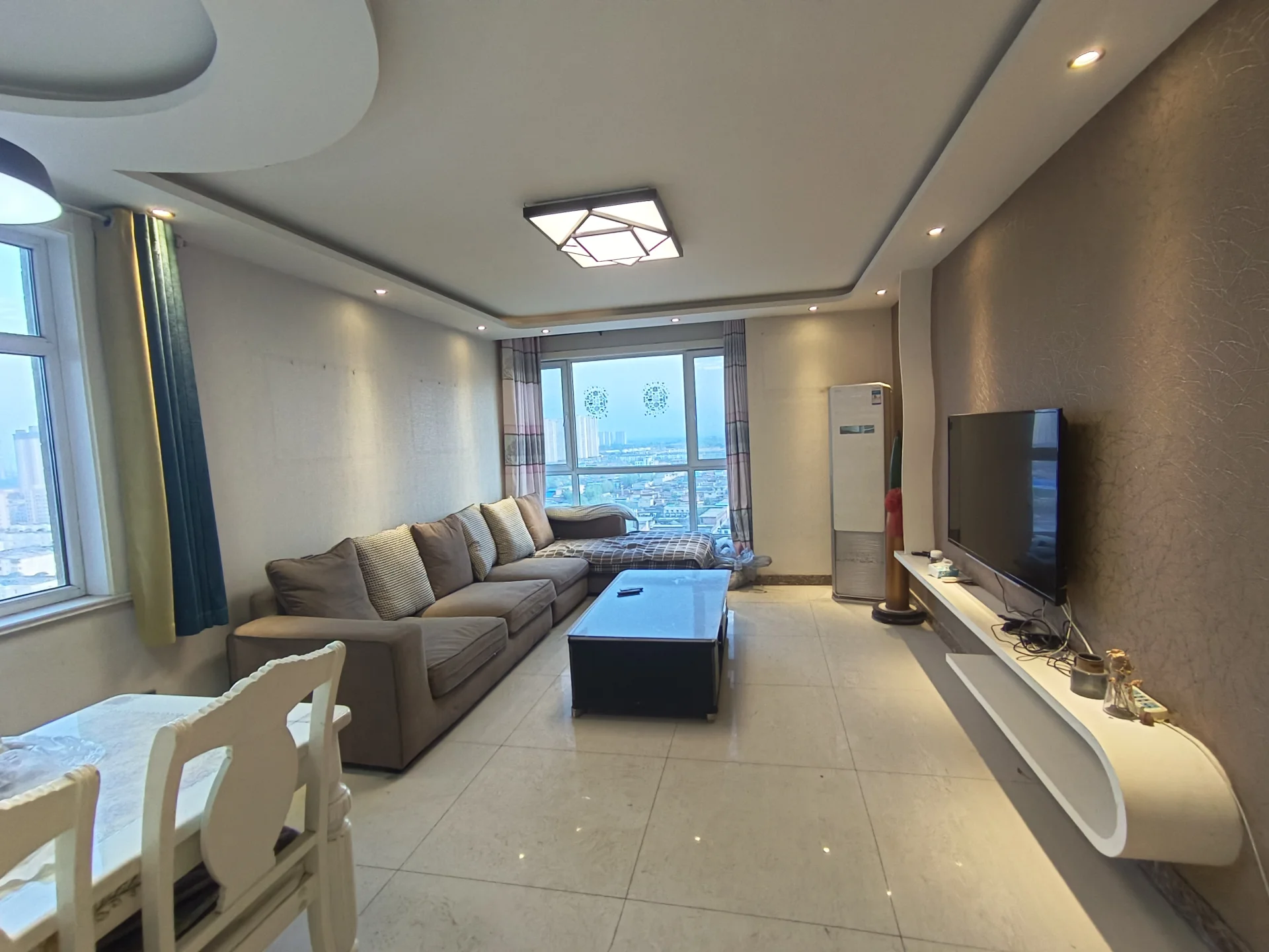 宽敞的客厅，白墙白地板，两套沙发分别位于左侧和中间，中间摆放着咖啡桌。电视挂在右侧墙面上。大窗户提供城市景色。客厅内有两把椅子作为 dining area，墙上挂着花瓶和时钟。电视旁边放着遥控器。关键字:宽敞 白墙 沙发 咖啡桌 电视 城市景色 椅子 花瓶 时钟 遥控器