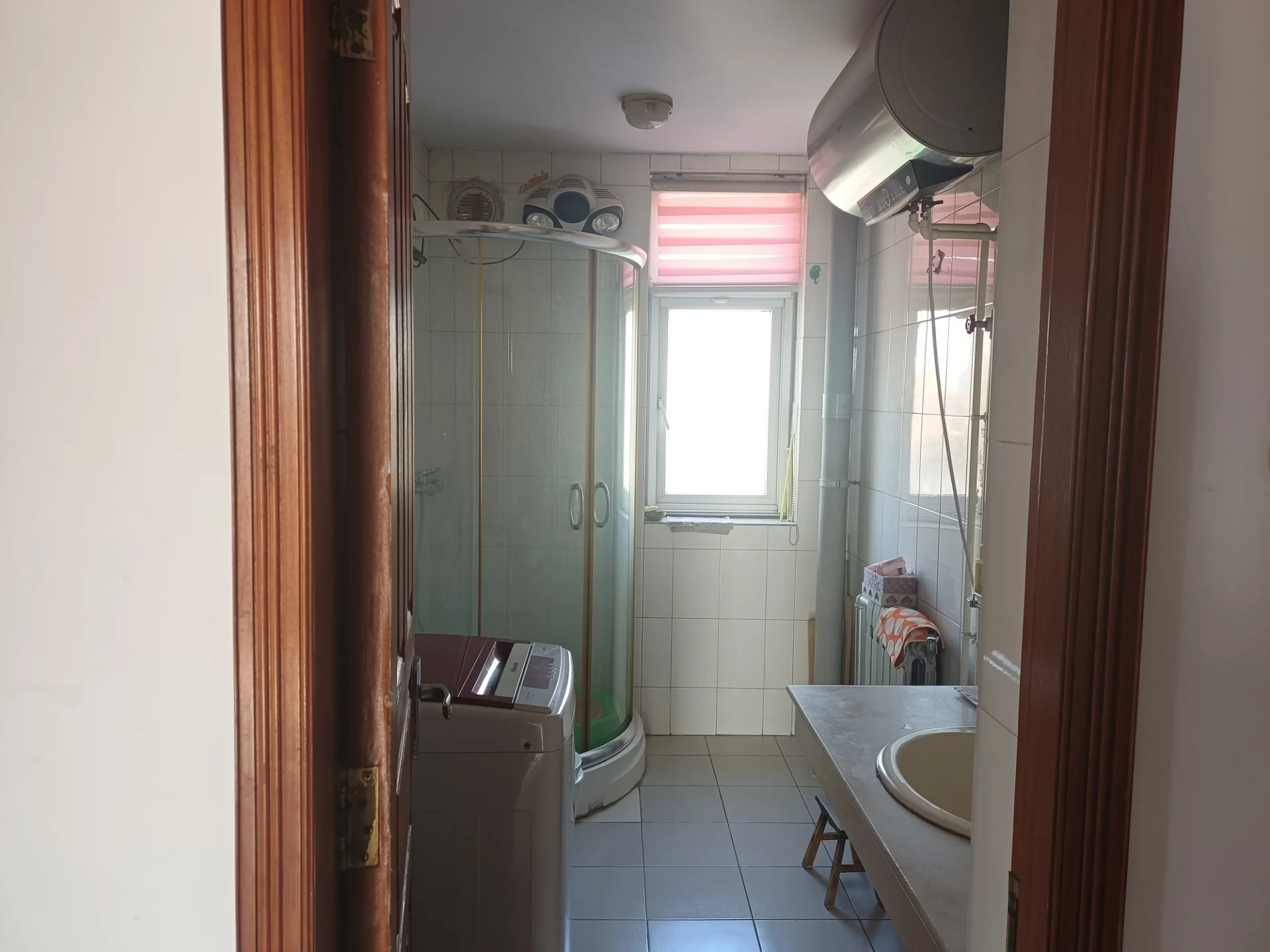 小而整洁的卫生间，白色调，配有洗手盆、马桶、镜子和淋浴。两扇窗户引入自然光，洗手盆上方有储物柜，旁边摆放着一把椅子。整体氛围干净有序。关键字:小卫生间 白色调 洗手盆 马桶 镜子 淋浴 自然光 储物柜 椅子