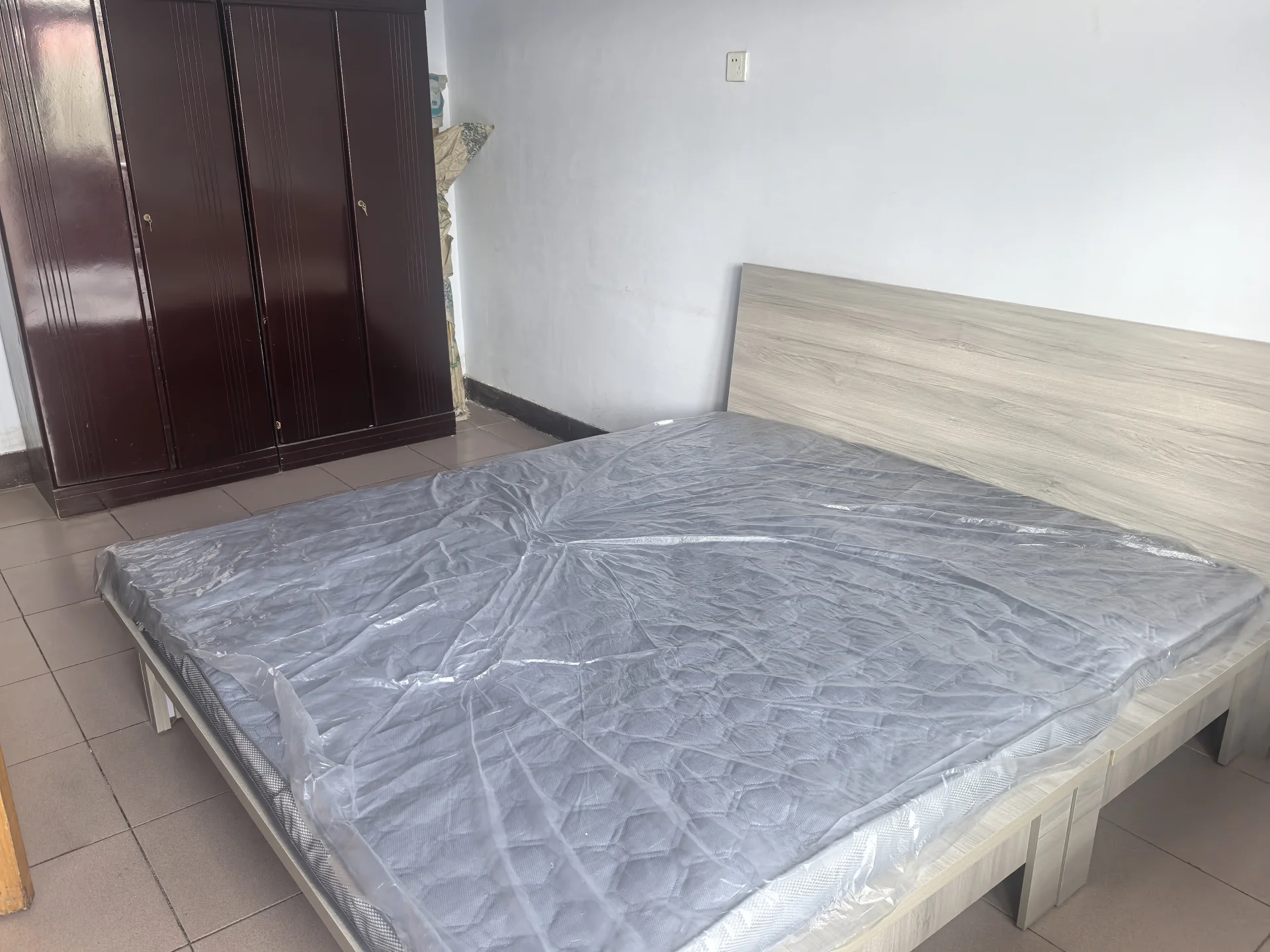 卧室中有一张大床，床上铺着塑料布和床垫，木制床头柜置于其后。两侧各有一只夜桌，房间显得空旷而简约，营造出宁静的氛围。关键字:卧室 大床 塑料布 床垫 木制床头柜 夜桌 空旷 简约 宁静