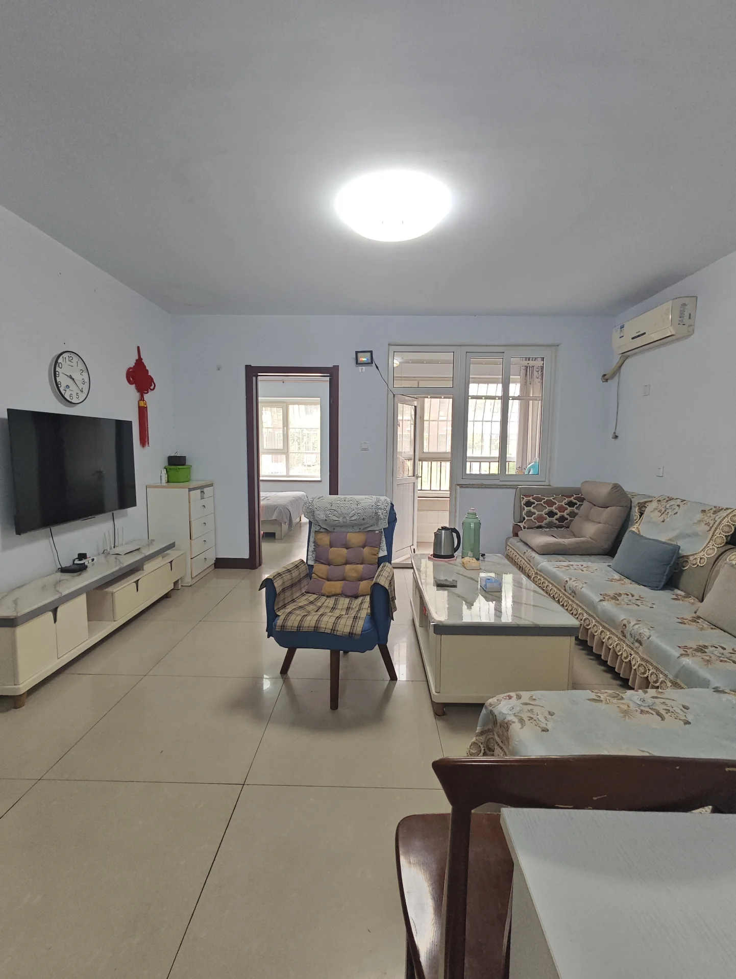 宽敞明亮的客厅，硬木地板，两侧各有一张沙发，中间摆放一把椅子。电视挂在左侧墙壁上，上方悬挂着一个时钟。客厅通向餐厅，配备餐桌和椅子。厨房紧邻餐厅，有冰箱和水槽。角落里摆放一盆植物，增添绿意。整体氛围宁静宜人，是放松或招待客人的理想之地。关键字:宽敞 明亮 硬木地板 沙发 椅子 电视 时钟 餐厅 餐桌椅 厨房 冰箱 水槽 植物