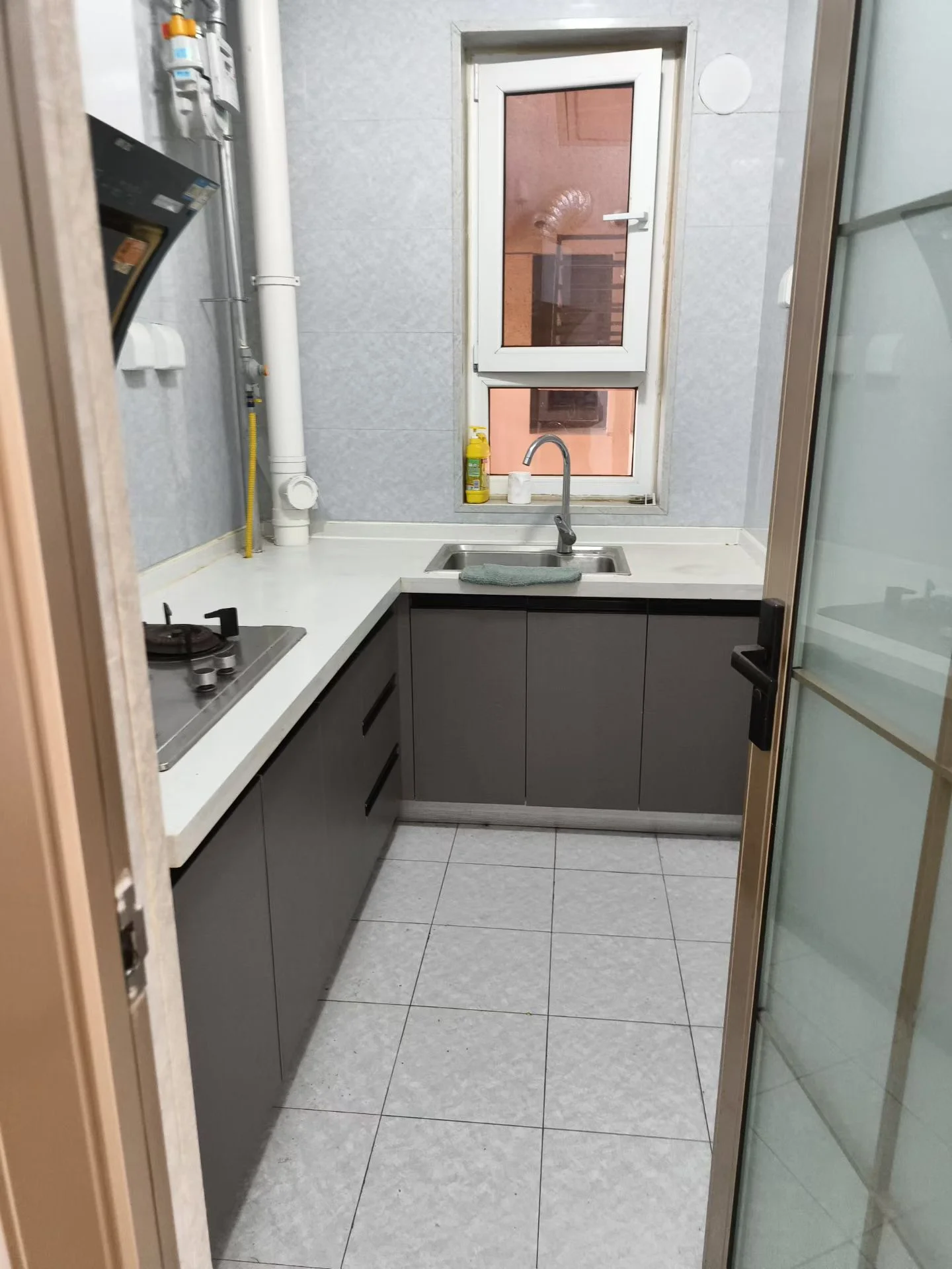 小而干净的厨房，白瓷砖地面和墙面，窗户位于水槽上方，自然光充足。橱柜下方用于储物，厨房空无一人，显得宁静而空旷。简约风格，明亮开阔。关键字:小厨房 白瓷砖 自然光 储物柜 简约风格