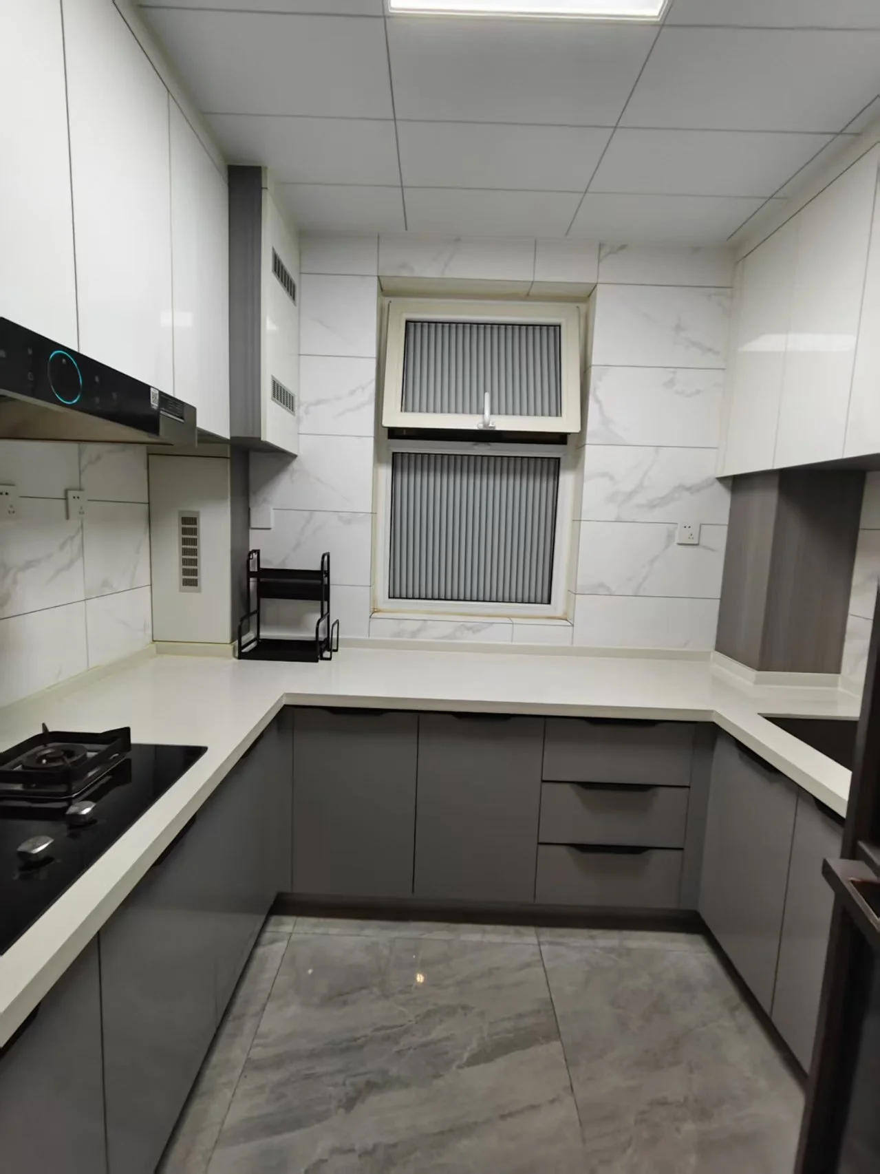 厨房整洁有序，白色橱柜与大理石台面搭配，灰色家电点缀。大窗户上方设有水槽，自然光充足。配备炉灶、烤箱、微波炉和冰箱。厨房用具如刀叉整齐摆放，营造出宁静舒适的氛围，是烹饪和家庭聚会的理想场所。关键字:厨房 整洁 白色橱柜 大理石台面 灰色家电 大窗户 自然光 炉灶 烤箱 微波炉 冰箱