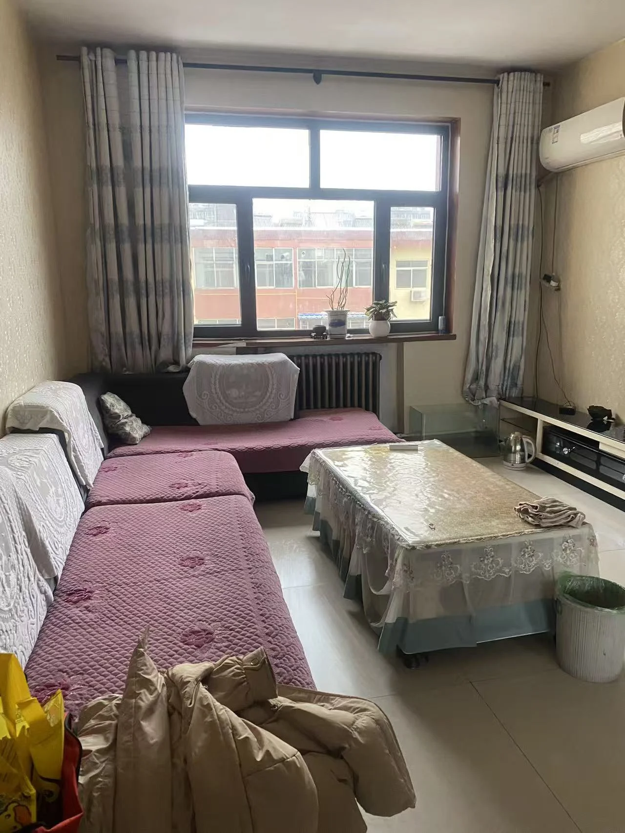 温馨舒适的客厅，白色瓷砖地面，沙发左角，床右侧，电视悬挂在床上方。两盆植物点缀其间，餐桌椅环绕，桌上三杯饮品暗示放松时光。背景冰箱可见，开放式厨房与客厅布局。关键字:小客厅 温馨舒适 白色瓷砖 沙发 床 电视 植物 餐桌椅 饮品 开放式布局