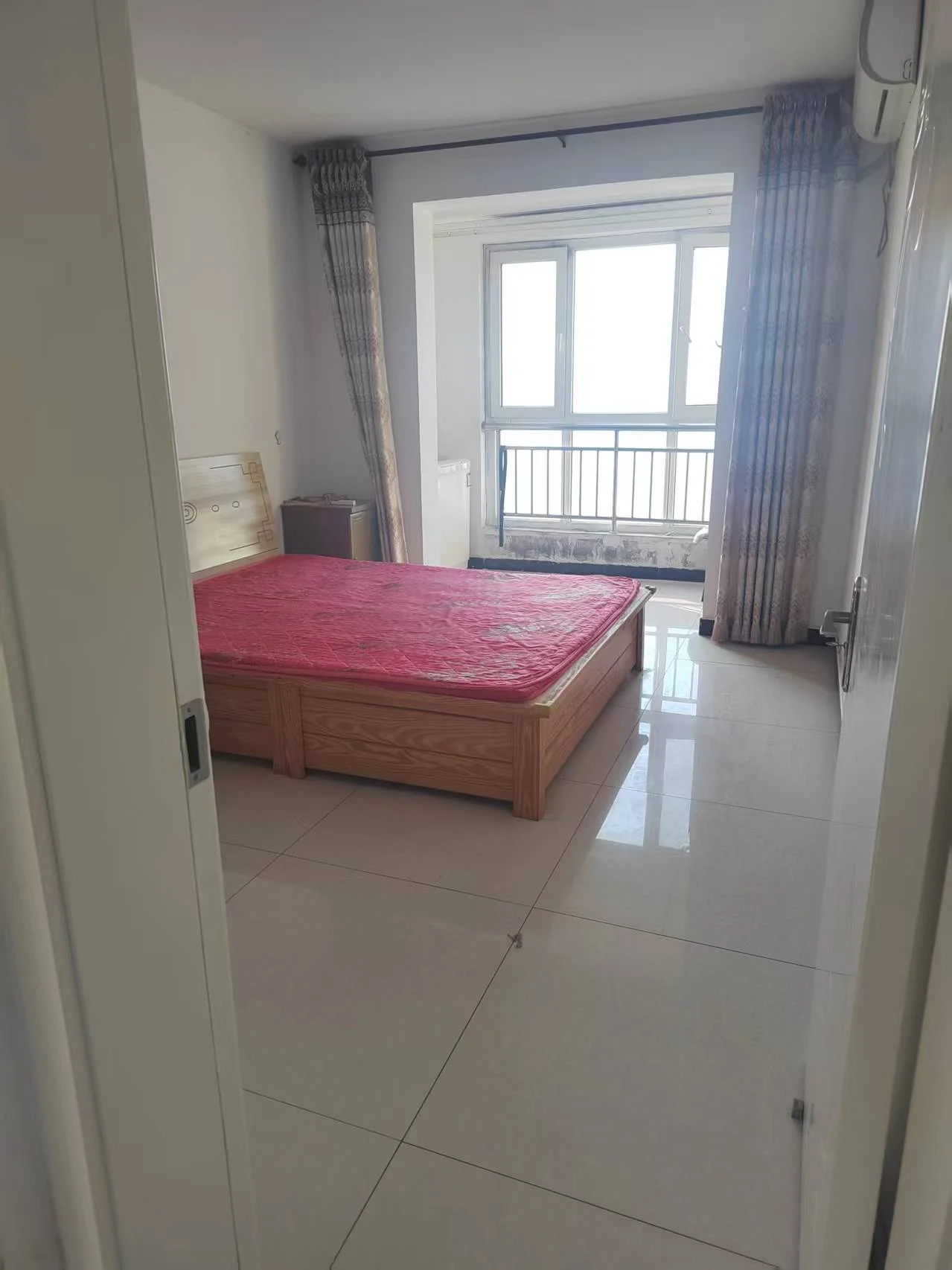 这间小卧室干净整洁，白色瓷砖地面和墙壁。一张红色的床置于房间中央，窗外是户外露台的景色。目前房间显得空旷无人，氛围宁静祥和，没有杂乱或喧嚣。简约的建筑设计，白色墙面和地板营造出干净利落的视觉效果。关键字:小卧室 白色瓷砖 红色床 露台 宁静祥和 简约设计