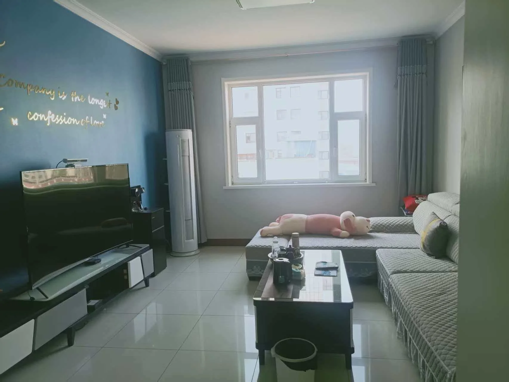 宽敞明亮的客厅，白色墙面，电视和沙发分置两侧，中央摆放咖啡桌和椅子。自然光透过窗户洒入，营造温馨氛围。餐桌附近有碗和杯子，沙发旁摆设两个泰迪熊，增添舒适感。关键字:客厅 白色墙面 电视 沙发 咖啡桌 椅子 自然光 窗户 餐桌 碗 杯子 泰迪熊