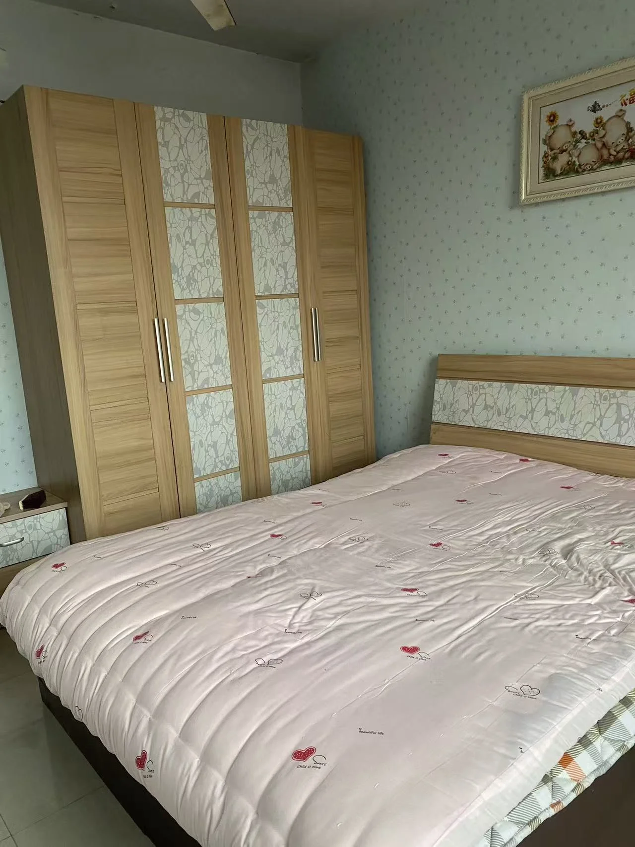 宽敞卧室，主景为大床，白色调搭配粉色床单，木制柜子上方摆放，营造宁静舒适的氛围。关键字:卧室 大床 白色调 粉色床单 木制柜子 宁静舒适