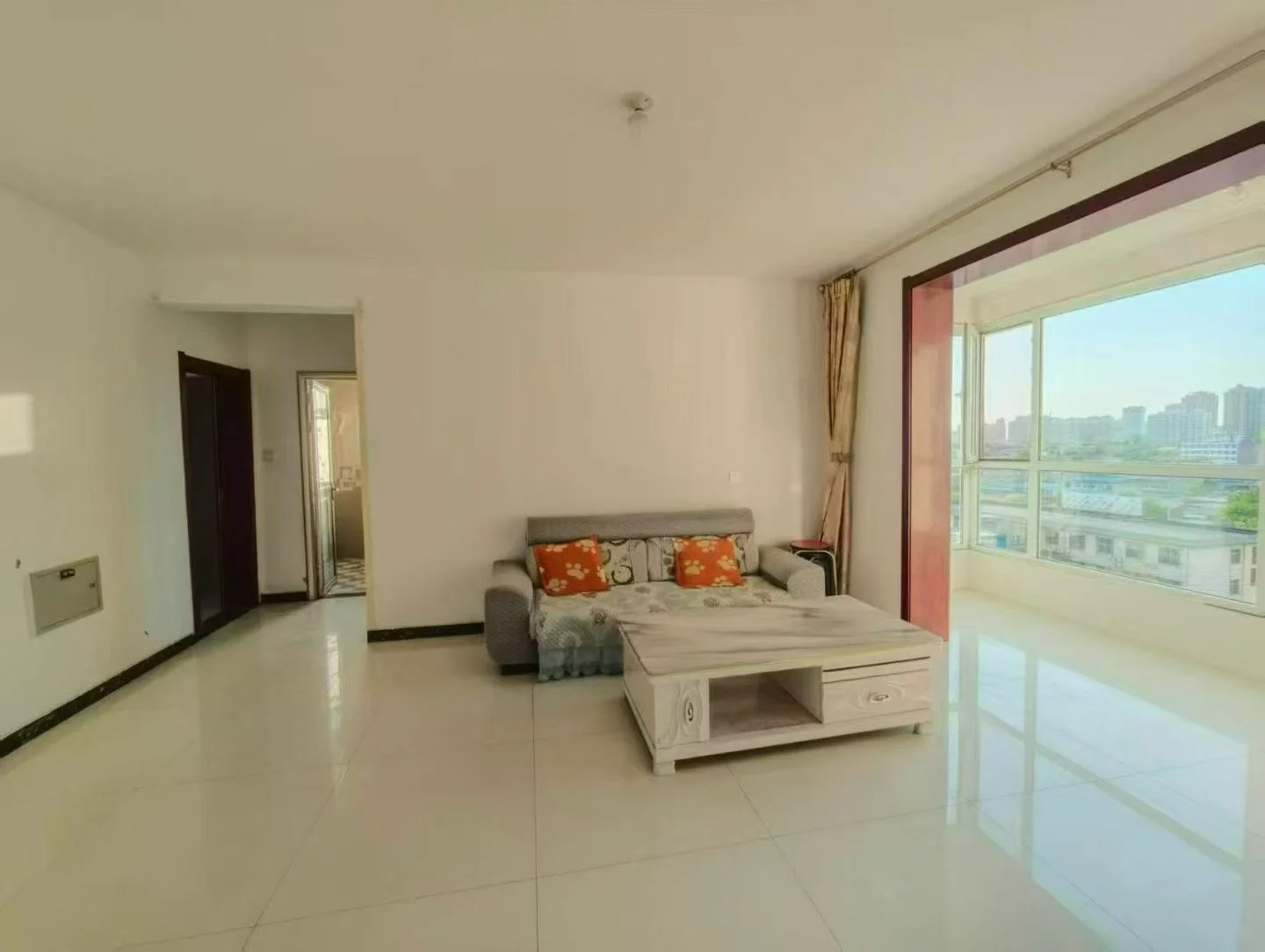 宽敞明亮的客厅，白色墙面，中置沙发配咖啡桌，窗大采光好，温馨舒适。沙发靠枕、花瓶点缀增添美感。开放式布局，背景可见厨房。关键字:宽敞 明亮 白色墙面 沙发 咖啡桌 大窗户 自然光 温馨舒适 靠枕 花瓶 开放式布局 厨房