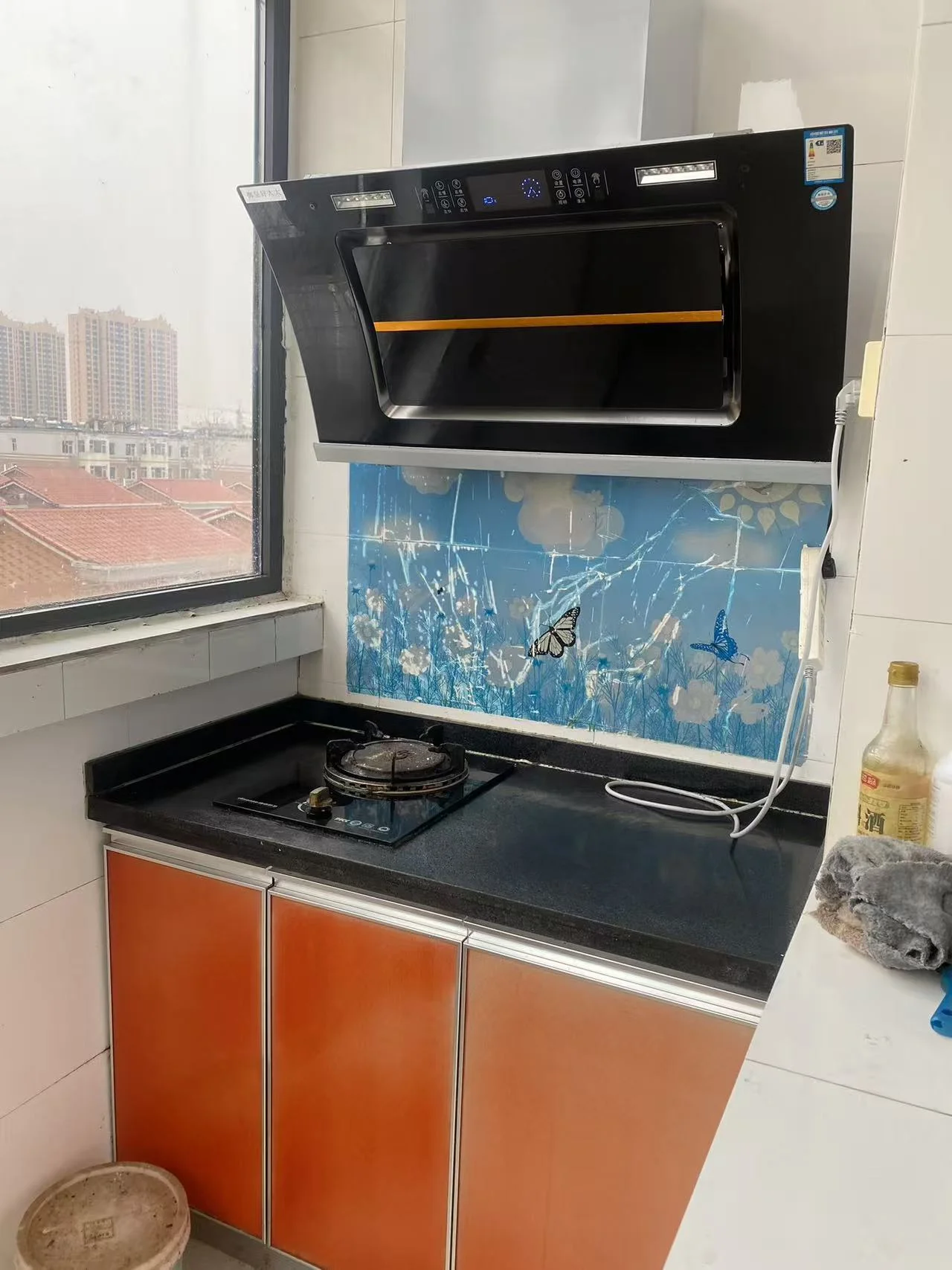 厨房内有烤箱和炉灶，烤箱为黑色，炉灶设计为蓝色和白色。两个水槽，其中一个在烤箱下方。上方有窗户，自然光充足。厨房整洁有序，水瓶、碗、勺子等物品整齐摆放。背景可见冰箱。整体氛围宁静宜人，是烹饪和用餐的理想空间。关键字:厨房 烤箱 炉灶 水槽 窗户 自然光 整洁 冰箱