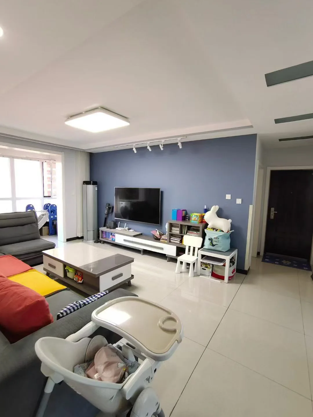 宽敞明亮的客厅，白色瓷砖地面，电视墙挂，沙发椅整齐排列，婴儿吊床增添温馨，大窗户采光自然，营造舒适氛围。关键字:客厅 白色瓷砖 电视墙挂 沙发椅 婴儿吊床 自然采光 舒适氛围