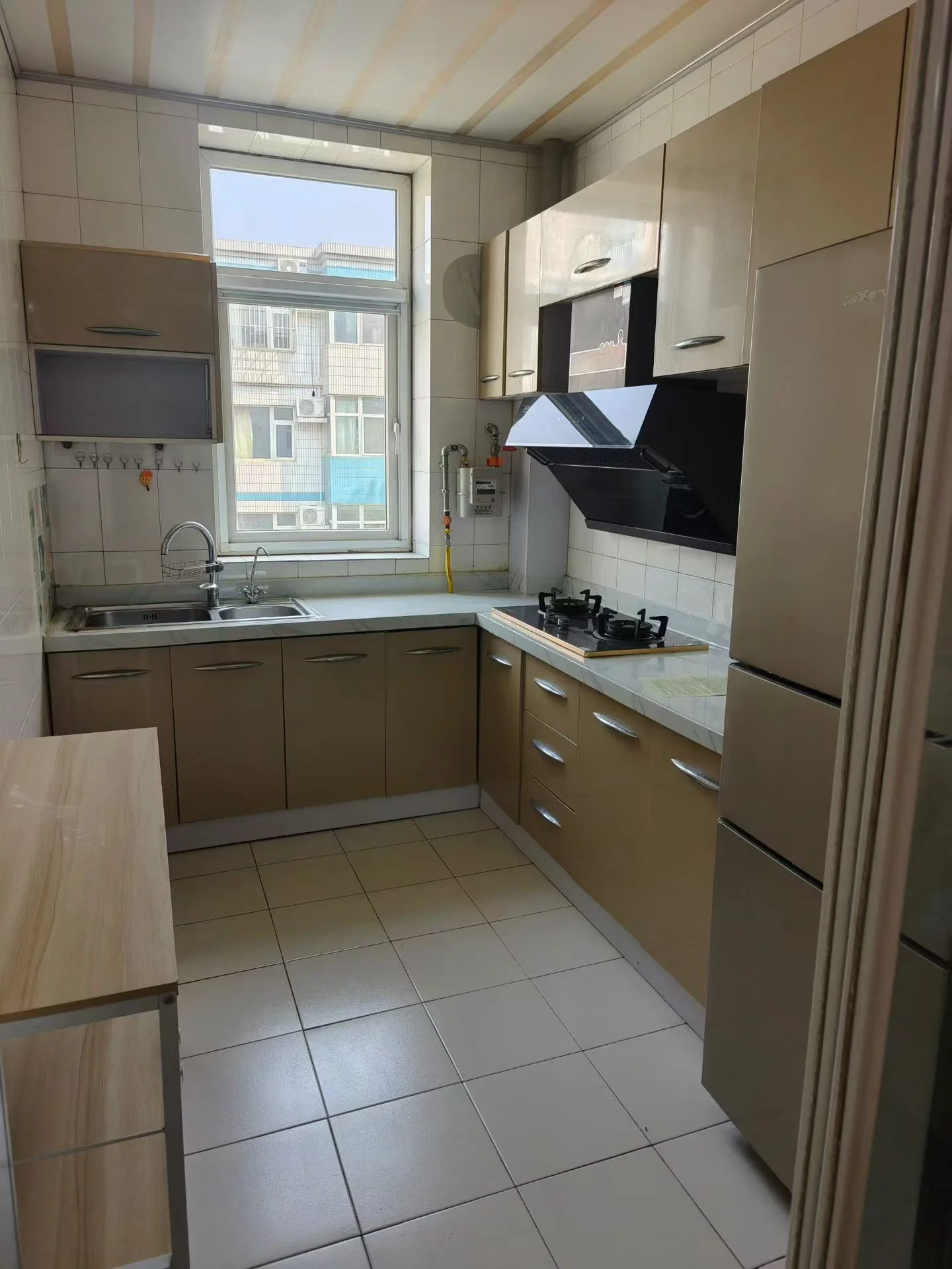 厨房干净整洁，白色瓷砖地面，白色橱柜和家电，自然光透过窗户洒入。不锈钢烤箱、水槽和冰箱等设备一应俱全，刀具和勺子整齐地摆放在 countertop 上。整体氛围宁静而简约，给人以舒适感。关键字:厨房 白色瓷砖 白色橱柜 自然光 不锈钢家电 刀具 勺子 宁静氛围