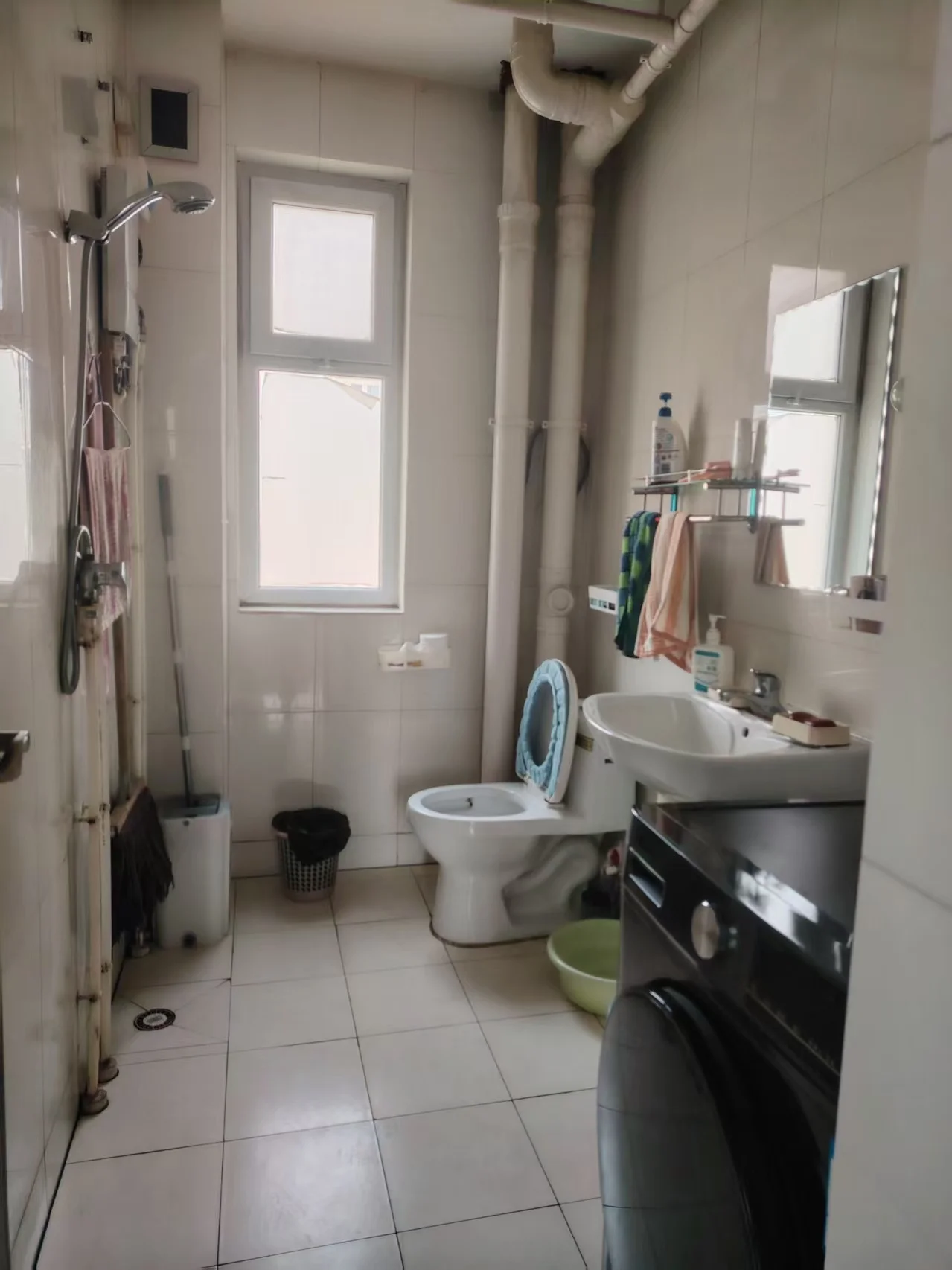 小而整洁的卫生间，白色瓷砖地面和墙面，配备马桶、洗手盆和淋浴。窗户引入自然光，台面放着几瓶瓶，显示频繁使用。整体氛围清新干净。关键字:小卫生间 白色瓷砖 马桶 洗手盆 淋浴 自然光 清洁