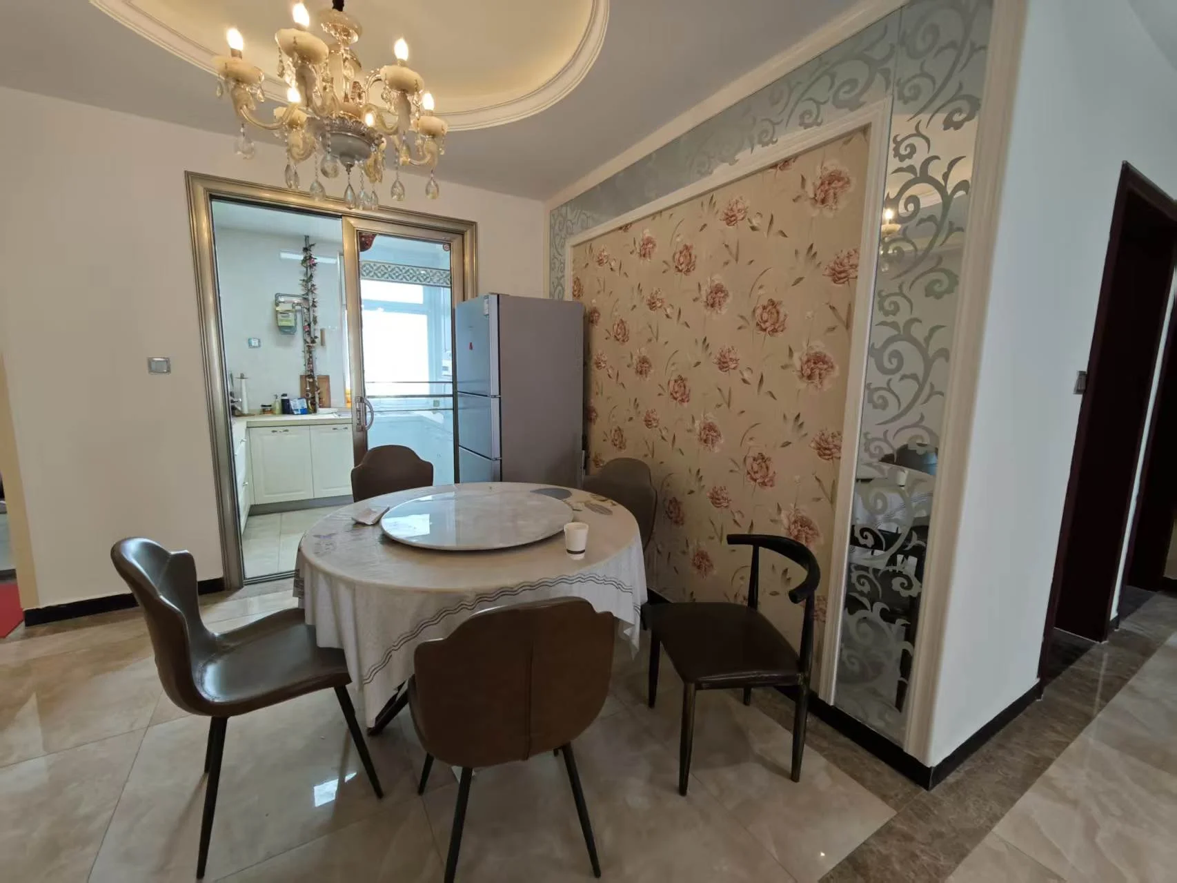 宽敞明亮的餐厅，大桌椅环绕，银器和餐具摆设整齐。上方吊灯增添优雅氛围。背景可见厨房，冰箱和水槽一应俱全。两扇窗户引入自然光，桌上一盆花增添装饰。关键字:宽敞 明亮 餐厅 大桌椅 银器 餐具 吊灯 优雅 厨房 冰箱 水槽 自然光 装饰