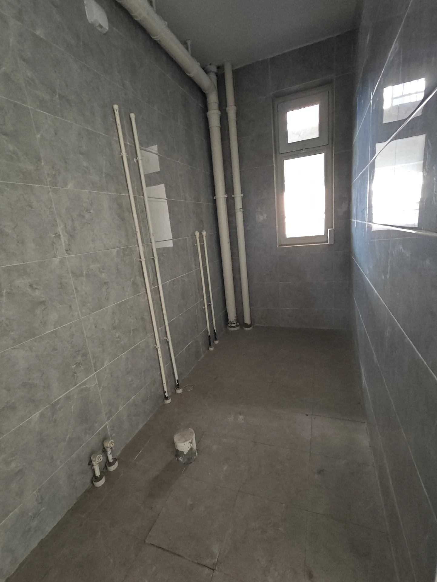 小浴室，白瓷砖墙面和地板，自然光透过窗户洒入。浴室显得正在施工或刚装修过，墙上有裸露的管道。整体氛围宁静而平静，无人在场。关键字:小浴室 白瓷砖 自然光 施工 装修 管道 宁静