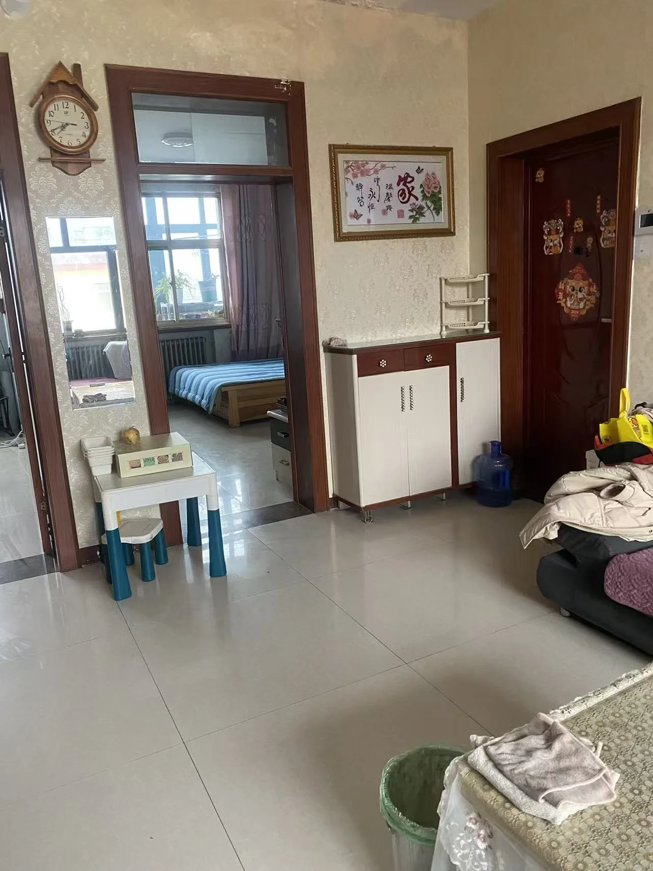 这是一间简洁明亮的亚洲小客厅，白色瓷砖地面和墙面营造出清新的氛围。墙上挂着一个时钟，增加实用性。房间内有两张床，一张靠近左侧，另一张靠近右侧。中间摆放着一张餐桌和一把椅子，旁边还有一间厨房ette配备Sink、冰箱和微波炉。左侧摆放着一盆植物，增添了一抹绿意。房间内几瓶饮料或杂物显露出一种宁静而亲切的氛围，突出了功能性和整洁。关键字:小客厅 亚洲风格 白色瓷砖 明亮 清新 时钟 两张床 餐桌 椅子 厨房ette Sink 冰箱 微波炉 植物 宁静氛围