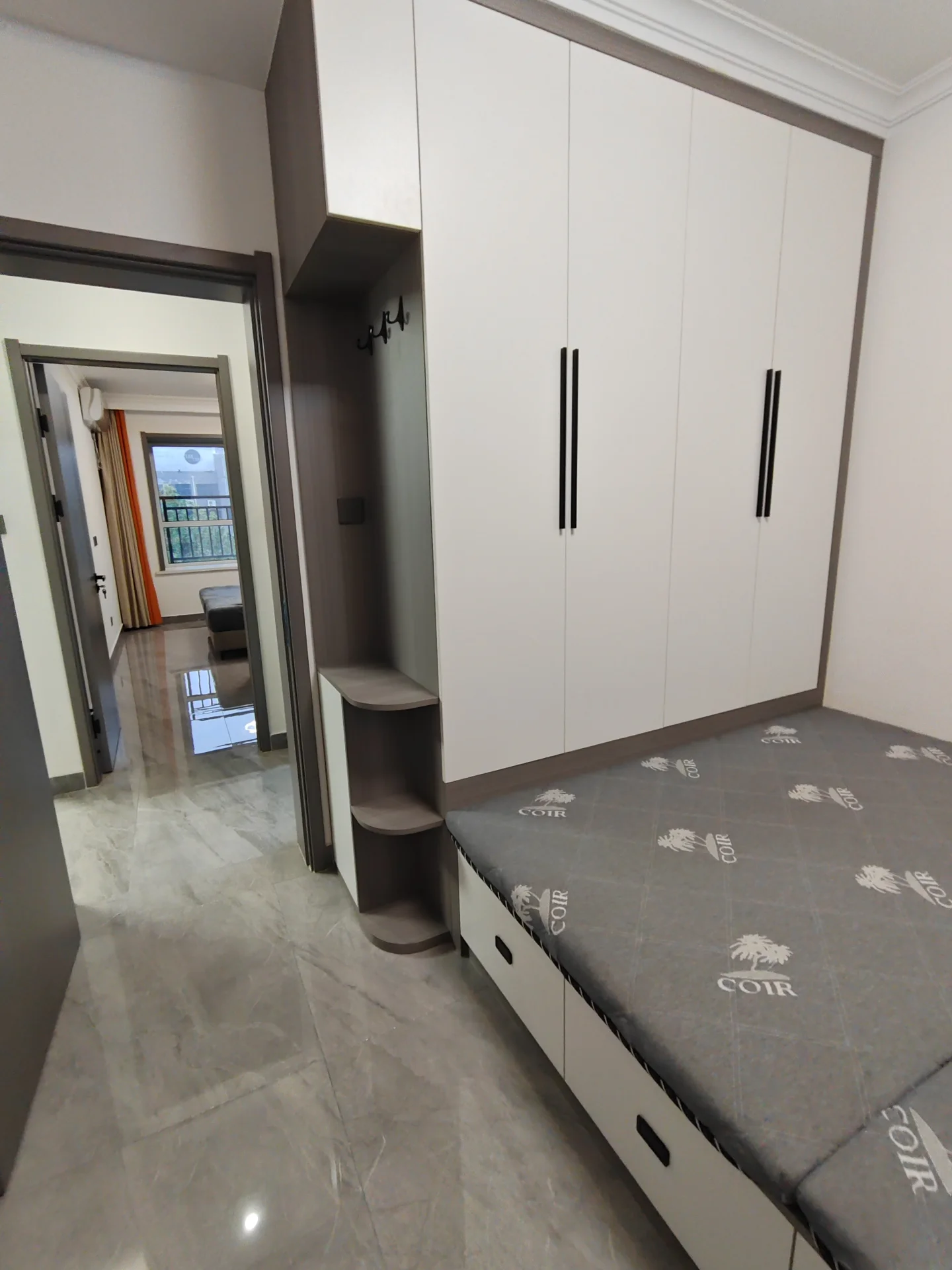 卧室以白色墙壁和灰色地板为主色调，宽敞的床仿佛置身酒店房间。床上方的两架书架上摆满了书籍，衣柜和镜子门通往另一间房间。整个空间显得宁静而祥和，没有丝毫活动的迹象。关键字:白色墙壁 灰色地板 大床 书架 衣柜 镜子门