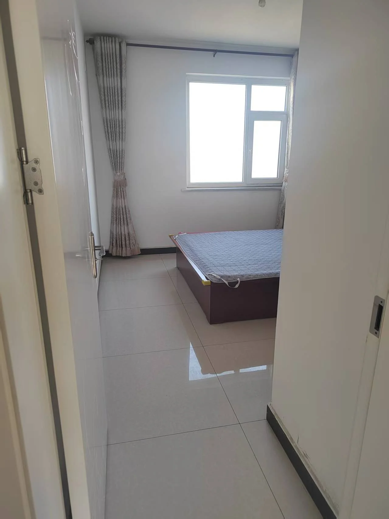 小卧室，白墙瓷砖地面，床铺蓝色被褥，窗户自然采光，窗帘增添装饰。关键字:小卧室 白墙 瓷砖地面 床铺 蓝色被褥 窗户 自然采光 窗帘