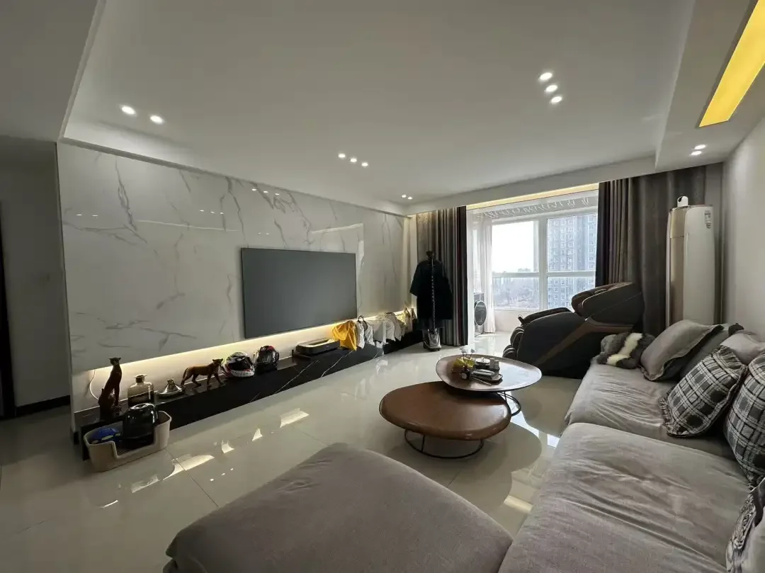 宽敞明亮的现代客厅，配备大屏幕电视、舒适的沙发和咖啡桌。厨房区域有冰箱、水槽和微波炉，旁边摆放着两把椅子。左侧有一张餐桌，装饰品如花瓶、书籍和酒瓶点缀其间。墙上挂着一个时钟，自然光从背景的两扇窗户洒入，营造出温馨舒适的氛围。关键字:宽敞 现代 客厅 大屏幕电视 沙发 咖啡桌 厨房 冰箱 水槽 微波炉 椅子 餐桌 装饰品 时钟 自然光