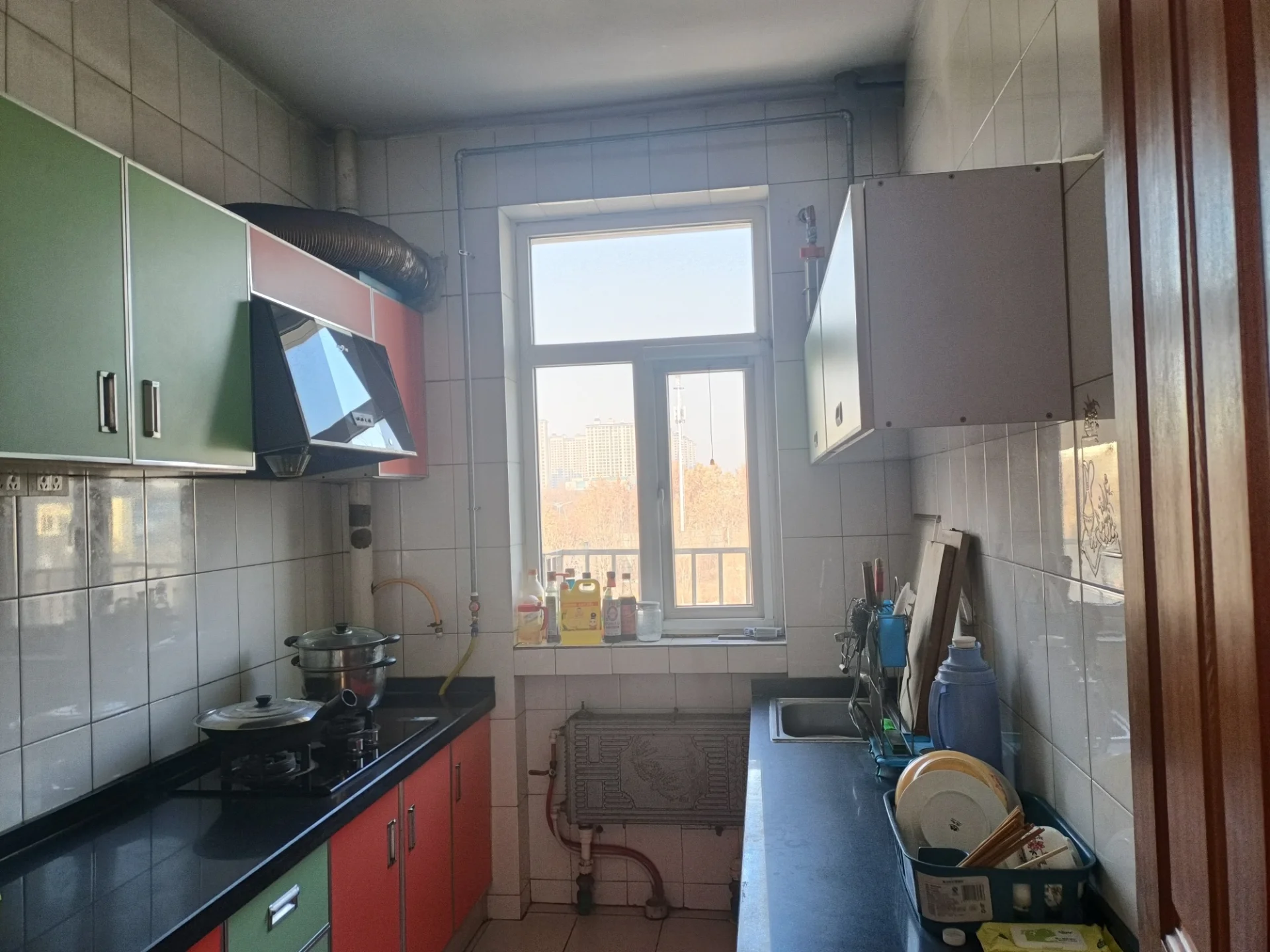 小厨房明亮整洁，白色瓷砖墙面和地面，窗户上方有水槽，自然光充足。厨房配备烤箱、炉灶、微波炉、冰箱等电器，以及刀具、勺子、碗、杯子和瓶子等餐具。 toaster和搅拌机等小家电增添便利。明亮的灯光营造温馨舒适的氛围，白色瓷砖使空间显得干净清新，实用且功能齐全。关键字:小厨房 明亮 白色瓷砖 自然光 烤箱 炉灶 微波炉 冰箱 刀具 勺子 碗 杯子 瓶子 toaster 搅拌机