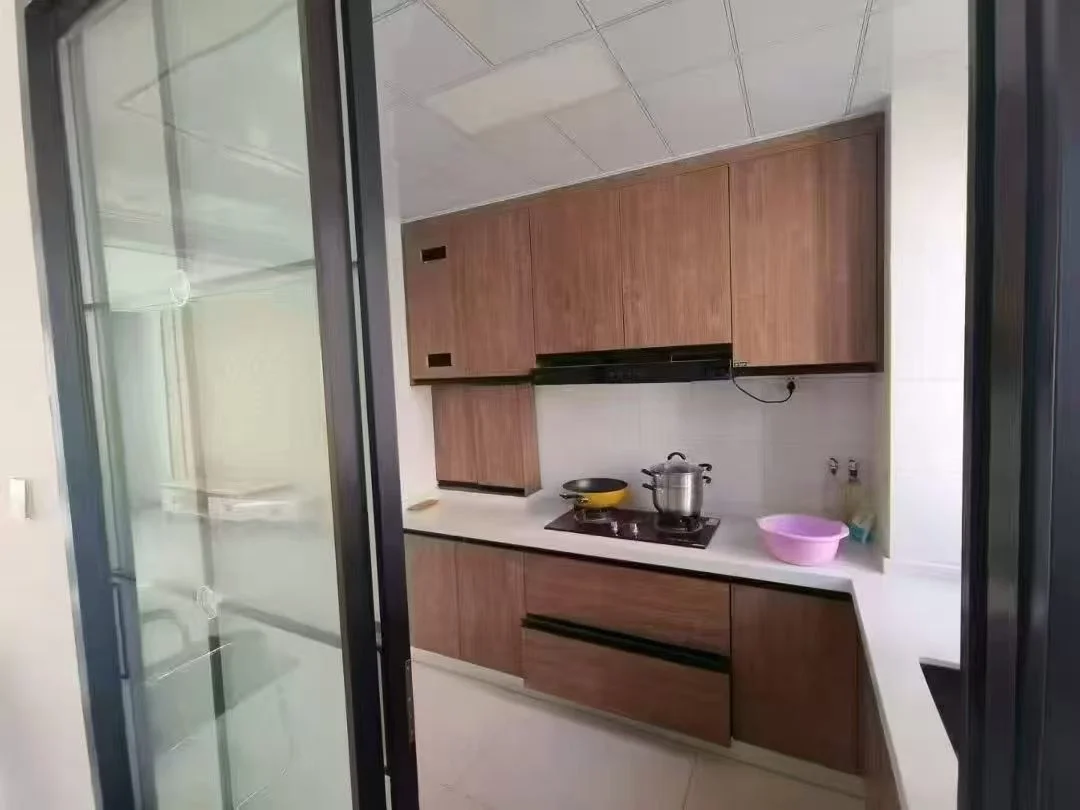 宽敞明亮的厨房，木质橱柜和白色台面，中央有水槽，一侧有烤箱，远处可见冰箱。厨房内摆放着碗、杯、勺和刀等物品，整体氛围宁静有序，充满功能性。关键字:宽敞 明亮 木质橱柜 白色台面 水槽 烤箱 冰箱 碗 杯 勺 刀