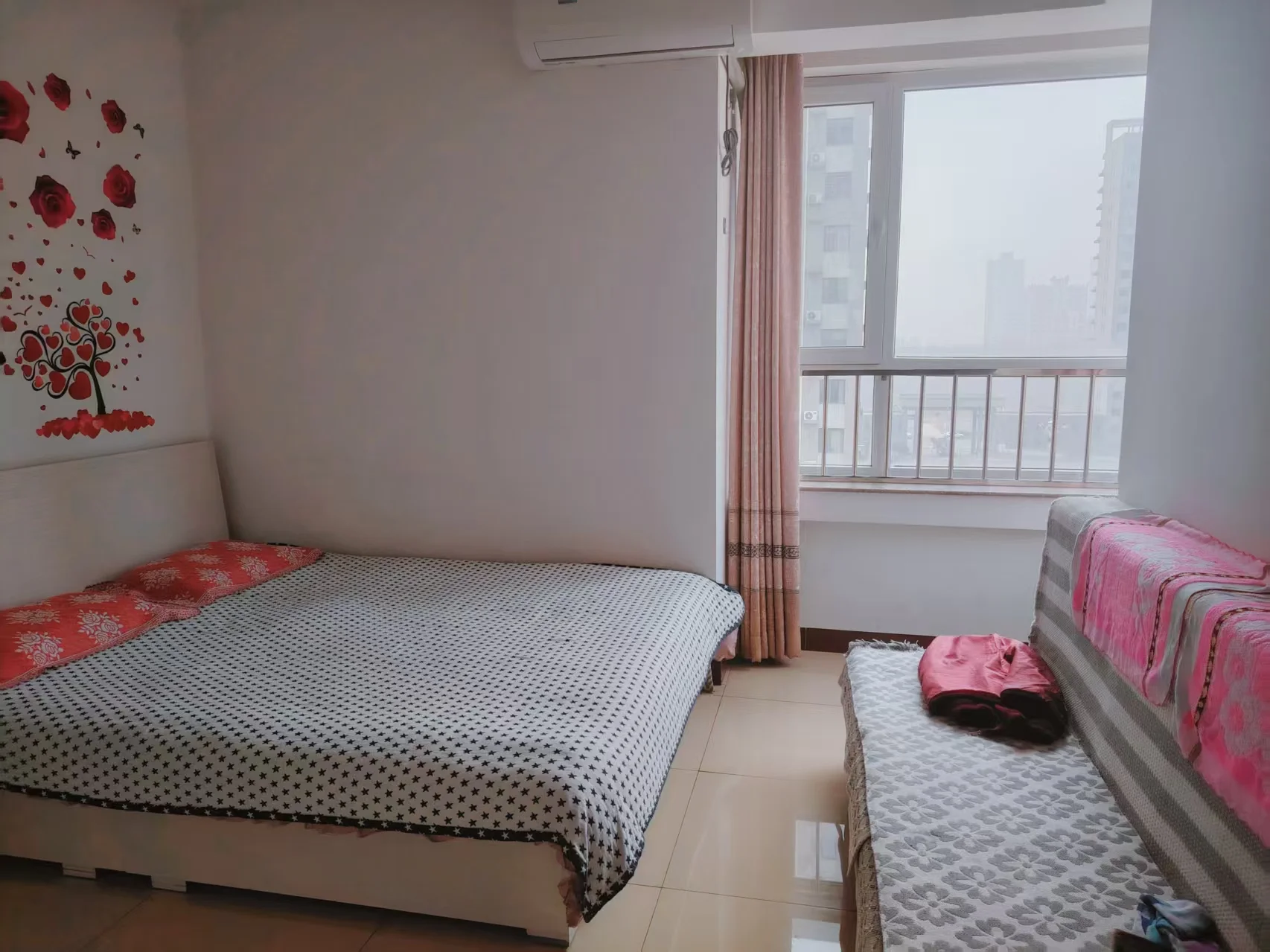 卧室里摆放了两张床，其中一张未整理。自然光透过窗户洒进房间，白色墙壁上点缀着红色装饰，增添了几分活力。一张粉色抱枕放在其中一张床上，角落里还摆放着一个泰迪熊。关键字:卧室 两张床 自然光 白色墙壁 红色装饰 粉色抱枕 泰迪熊