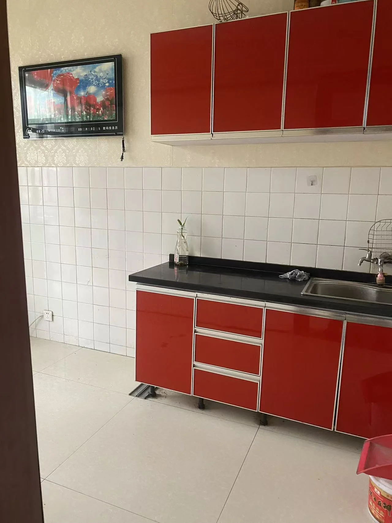 小厨房，红柜白墙，电视挂墙，冰箱靠窗，水槽旁有酒瓶，两侧各有花瓶。温馨舒适，色彩鲜明。关键字:小厨房 红柜 白墙 电视 冰箱 水槽 酒瓶 花瓶