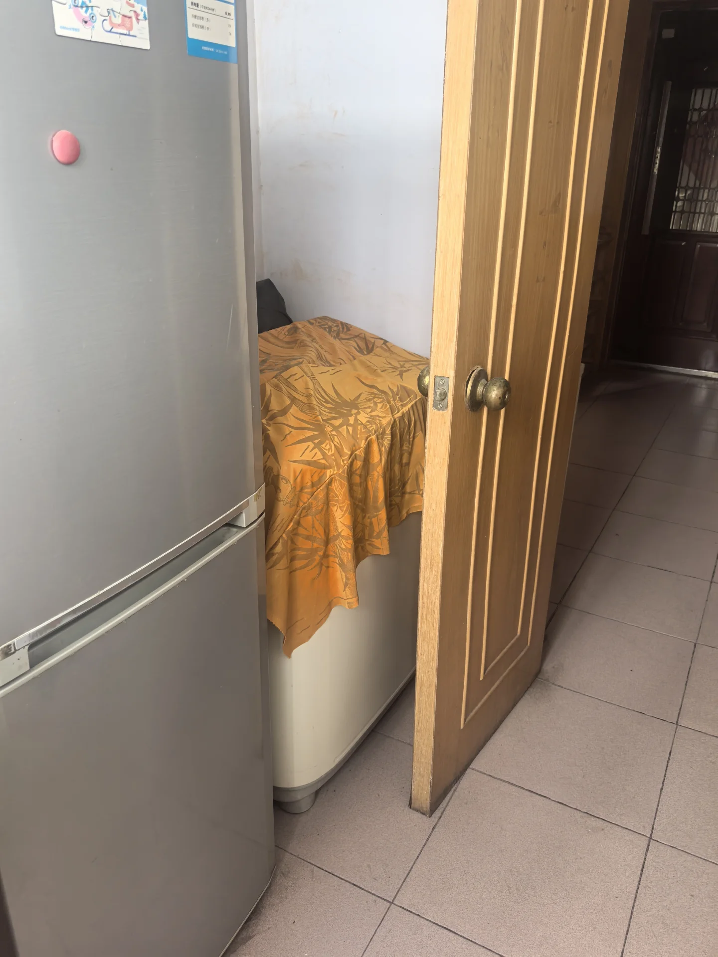 厨房左侧摆放着冰箱，右侧是通向另一间卧室的开放式门。卧室里铺有床单和被褥，显得整洁有序。关键字:厨房 冰箱 开放式门 卧室 床单 被褥 整洁有序