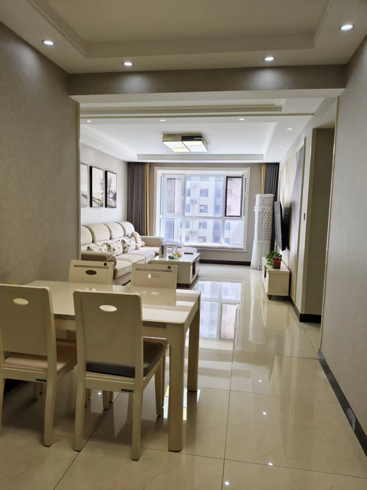 宽敞明亮的客厅，白色家具搭配，两侧各有餐桌椅，中央摆放另一张餐桌，沙发靠近一侧餐桌，绿植点缀四周。大窗户引入自然光，营造温馨舒适的氛围。墙上悬挂的电视增添了几分温馨感。关键字:宽敞 明亮 白色家具 餐桌椅 绿植 自然光 温馨氛围