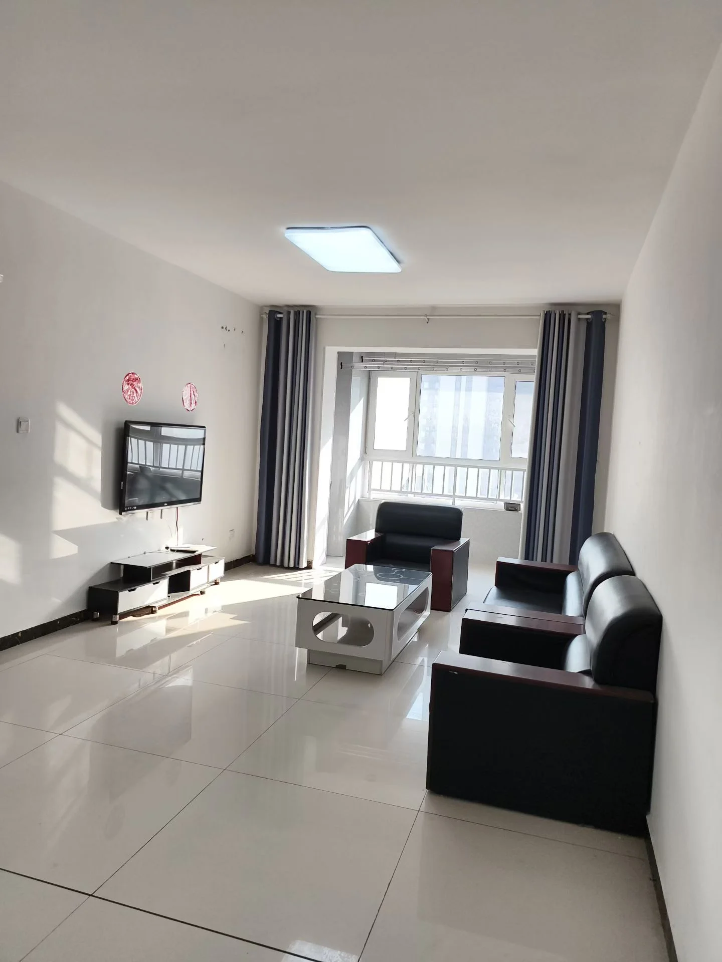 宽敞明亮的客厅，白色瓷砖地面，沙发居中摆放，面向电视，咖啡桌置于沙发与电视之间，两侧各有椅子，两扇大窗引入自然光，后方有餐桌，一盆植物增添绿意。关键字:宽敞 明亮 白色瓷砖 沙发 电视 咖啡桌 椅子 自然光 餐桌 绿植