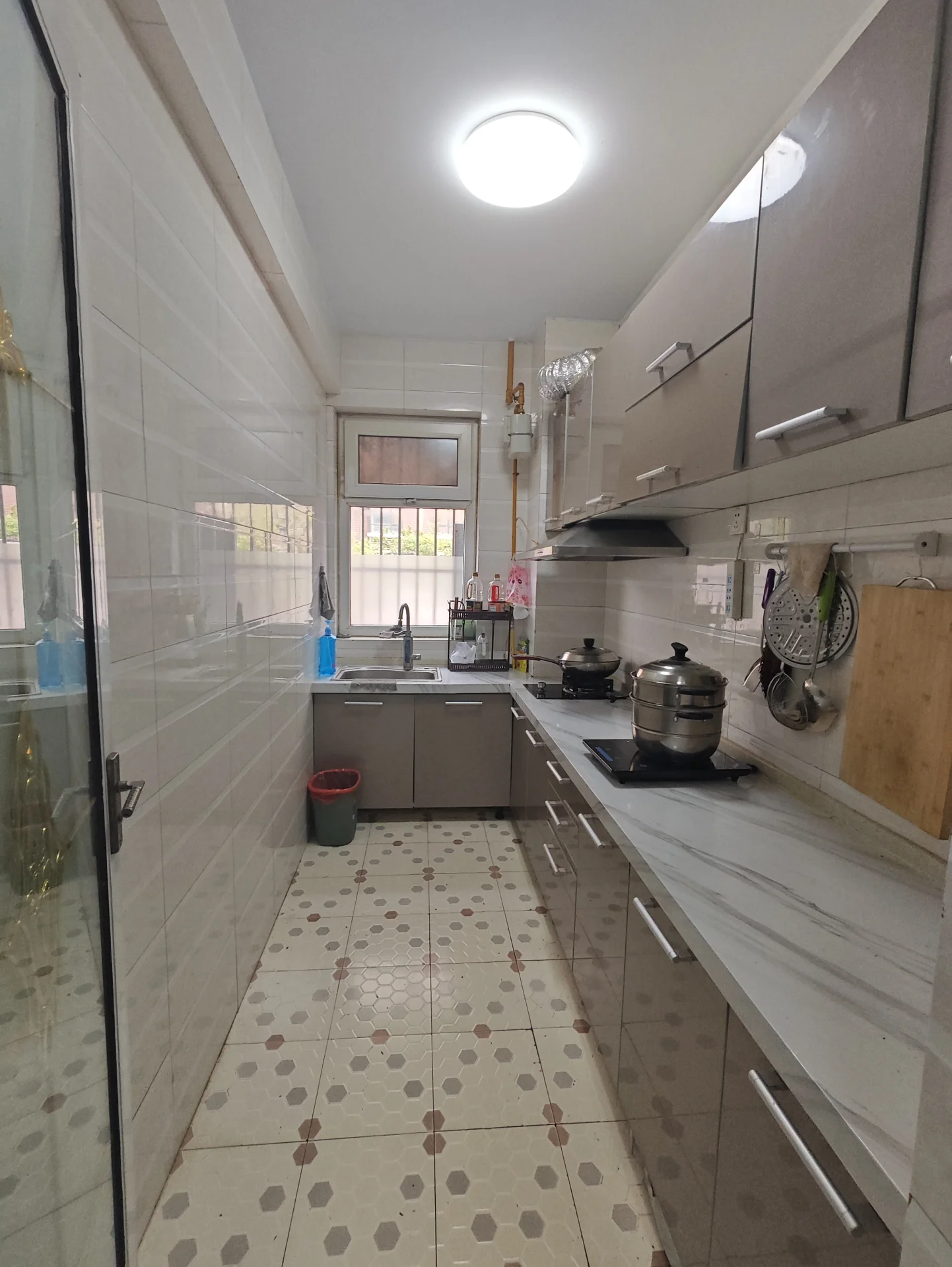 小而整洁的厨房，白色瓷砖地面，岛式设计中央，四面环绕橱柜。中央有水槽，厨具摆放在台面上。几瓶酒增添了几分温馨。厨房设备齐全，适合家庭或喜欢烹饪的个人使用。关键字:小厨房 白色瓷砖 岛式设计 橱柜 水槽 厨具 温馨氛围