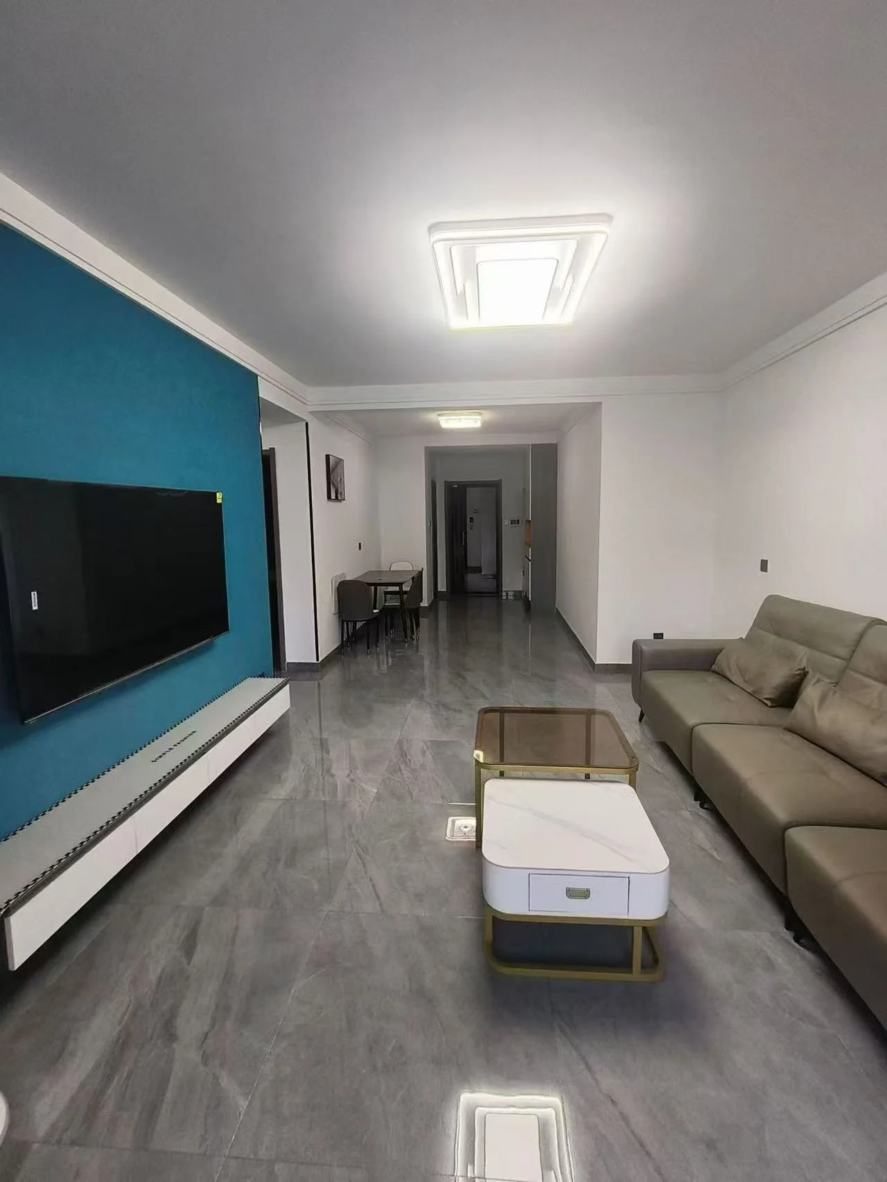 宽敞明亮的客厅，白色墙面，大屏幕电视墙上挂着，沙发背景，咖啡桌置于茶几与电视之间。旁边摆放两把椅子，左侧更有一把。客厅还有一张餐桌配几把椅子。遥控器放在桌上。自然光或人造光源营造出明亮氛围。关键字:客厅 白色墙面 大屏幕电视 沙发 咖啡桌 椅子 餐桌 明亮氛围