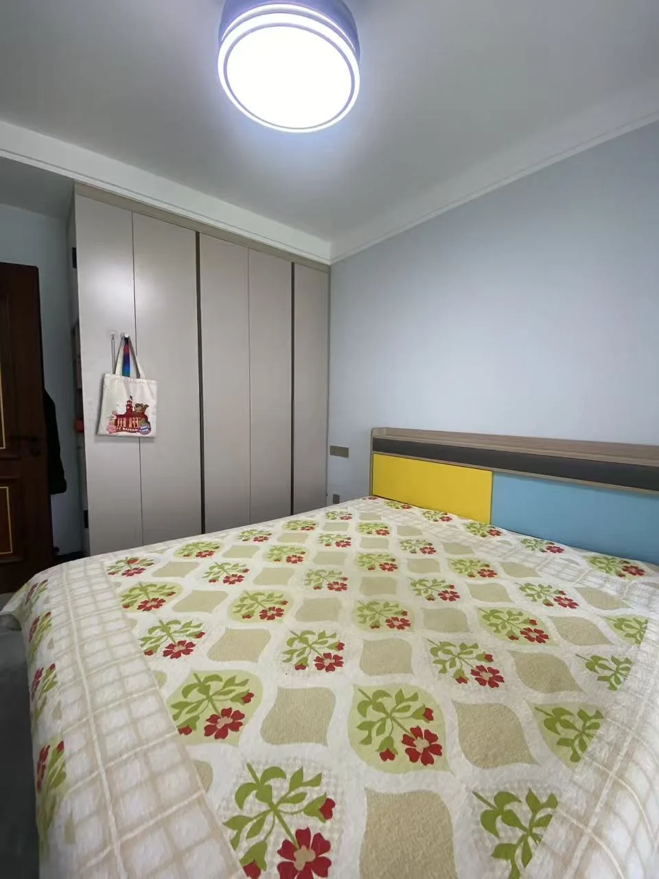 温馨舒适的卧室，大床居中，上方吊灯提供充足光线。黄色头板和蓝色墙面营造温暖氛围。两侧各有衣橱，床脚有长凳，床边放着手提包增添个人 touches。整体环境宁静舒适，适合放松休息。关键字:卧室 大床 吊灯 黄色头板 蓝色墙面 衣橱 长凳 手提包