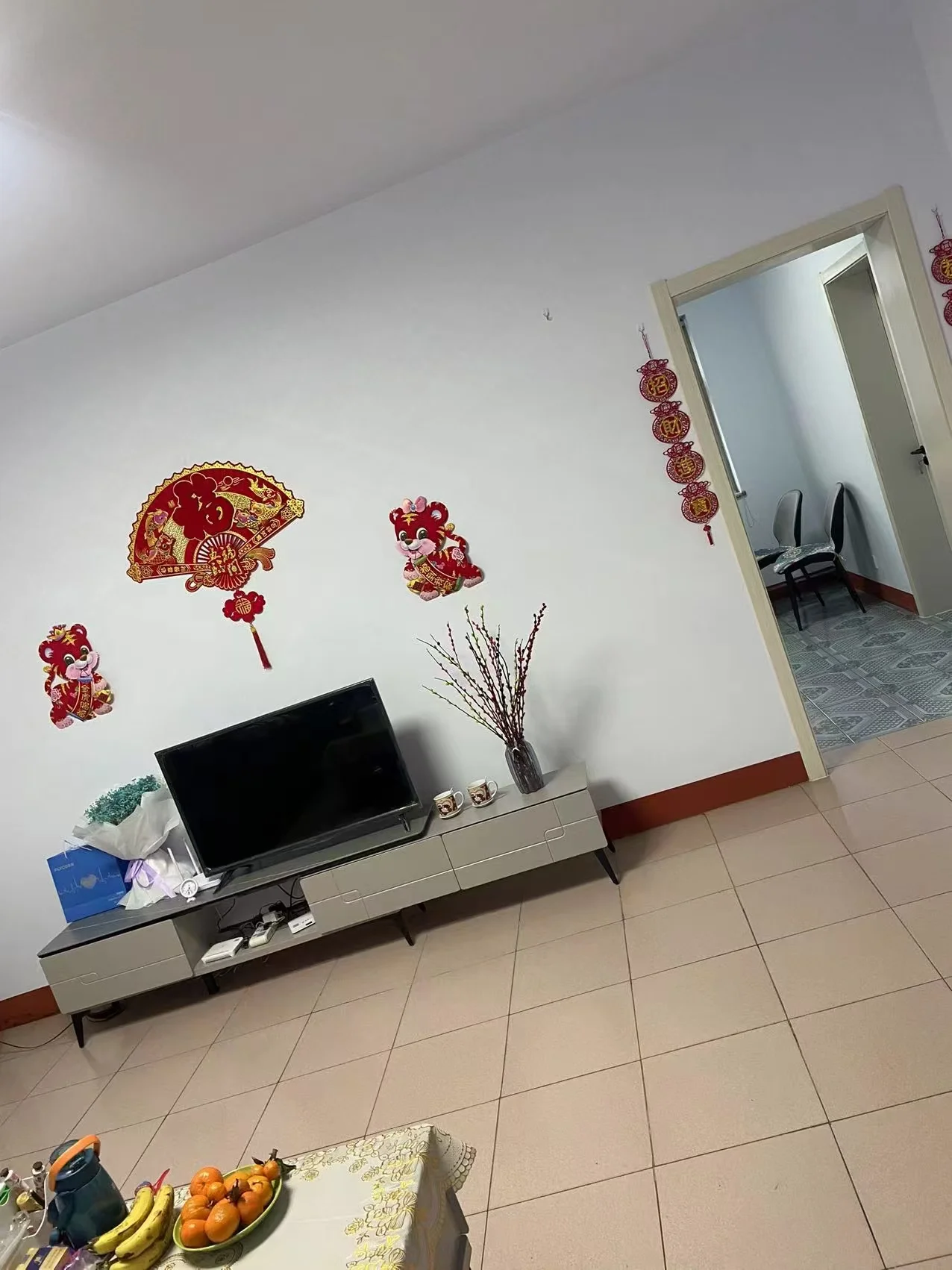 客厅空间，墙面为浅色调，配有电视和简约的电视柜。地面铺设米色瓷砖，整体光线明亮，呈现基础的居住环境。