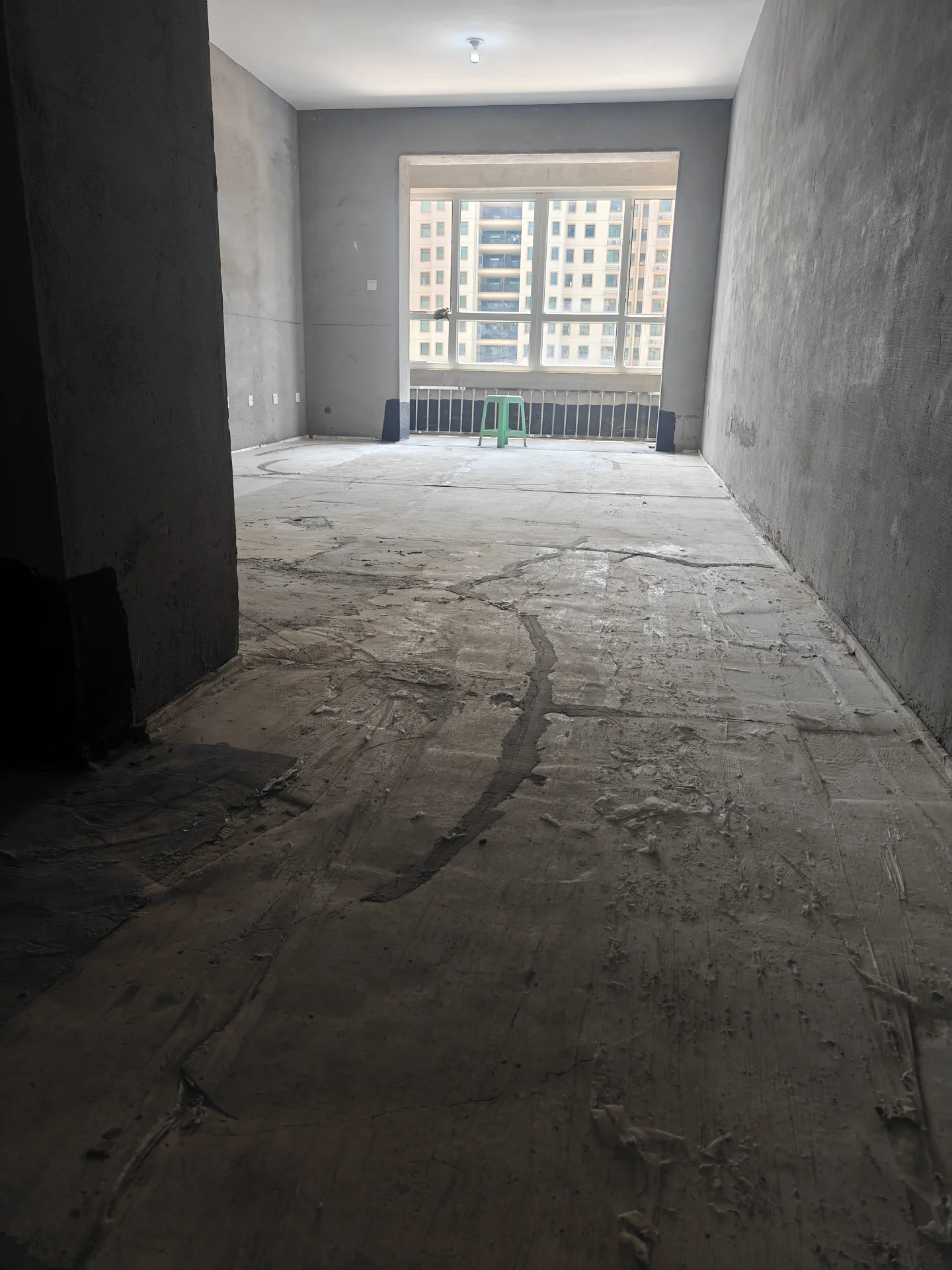 一个具有工业或原始风格的室内空间，以裸露的混凝土墙面和粗糙的地面为主，采光良好，背景可见外部的窗户和城市建筑的景象。