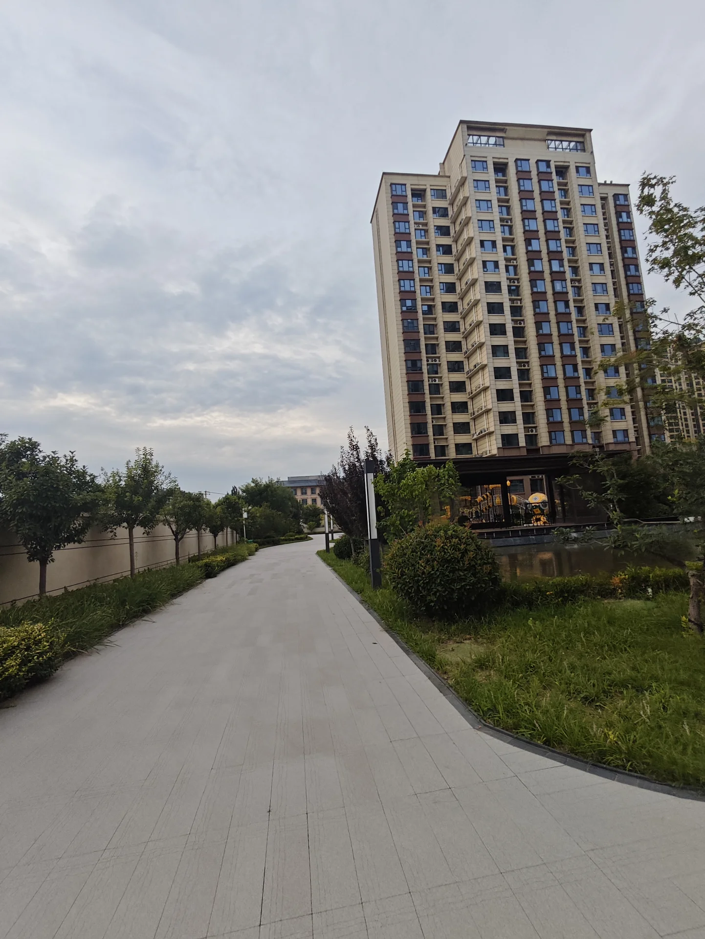 一栋多层现代风格的住宅楼矗立在前景，建筑立面采用浅色调，周围环绕着修剪整齐的绿化带和树木，整体呈现出一种宁静、宜居的社区氛围。