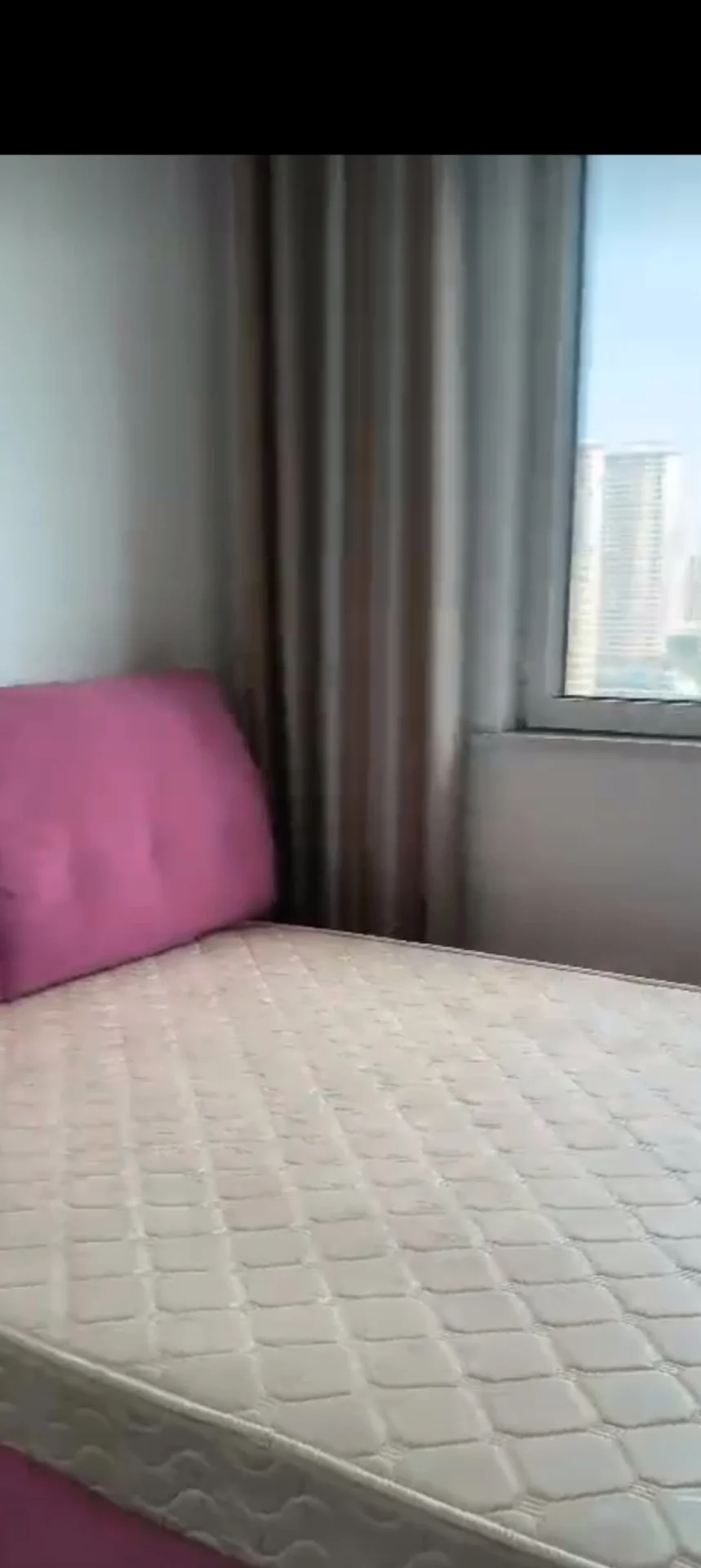 卧室场景，床铺铺着米色或浅色格纹床品，床头靠着粉色软包床头板，窗户被米色或棕色百叶窗遮挡，光线柔和，整体呈现温馨、舒适的卧室氛围。