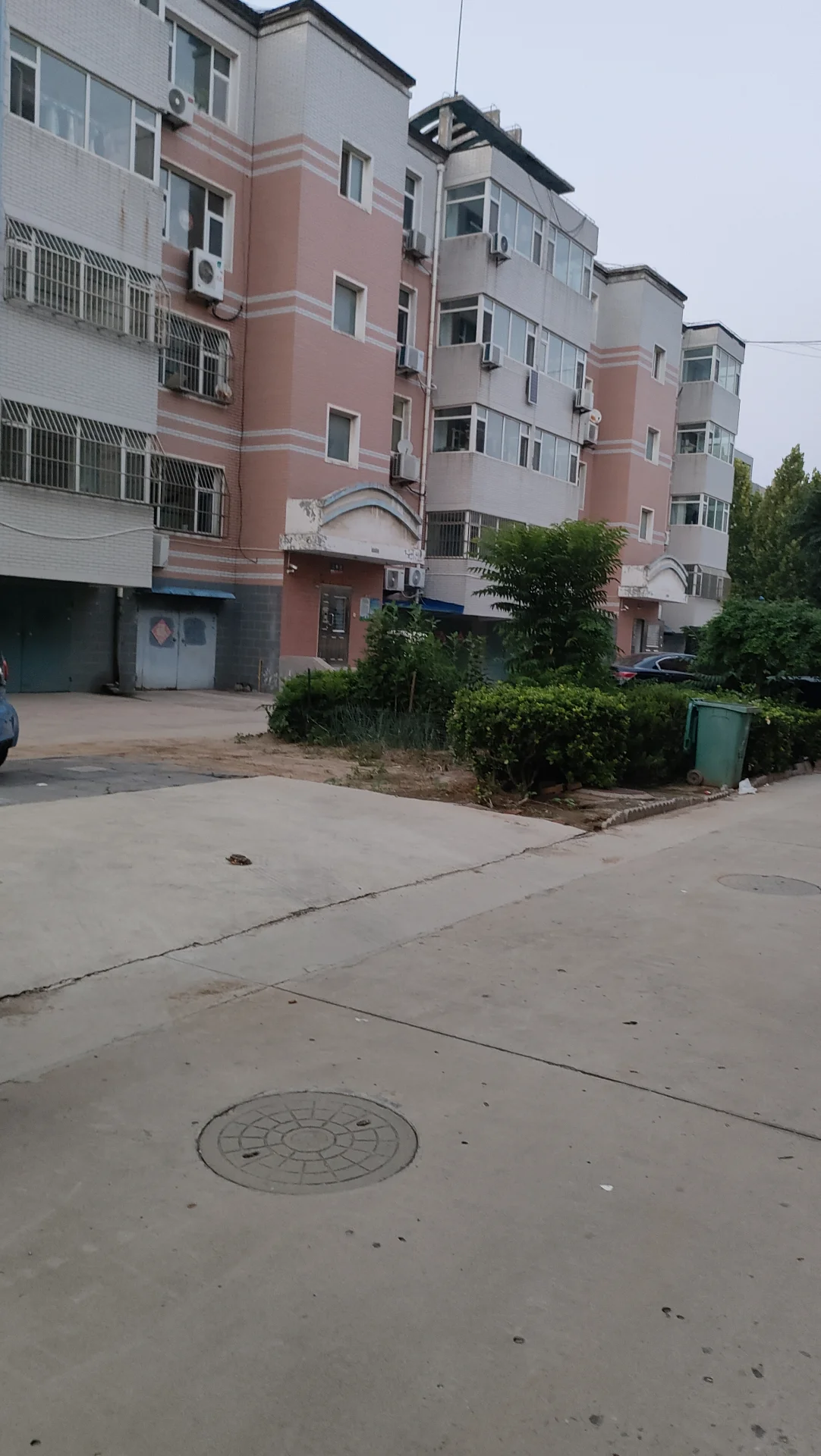 一栋多层住宅楼的外部立面，建筑主体呈现粉色和白色相结合的色调，设有多个窗户和阳台。建筑前方有低矮的绿植和人行道，整体呈现社区住宅的外部环境氛围。