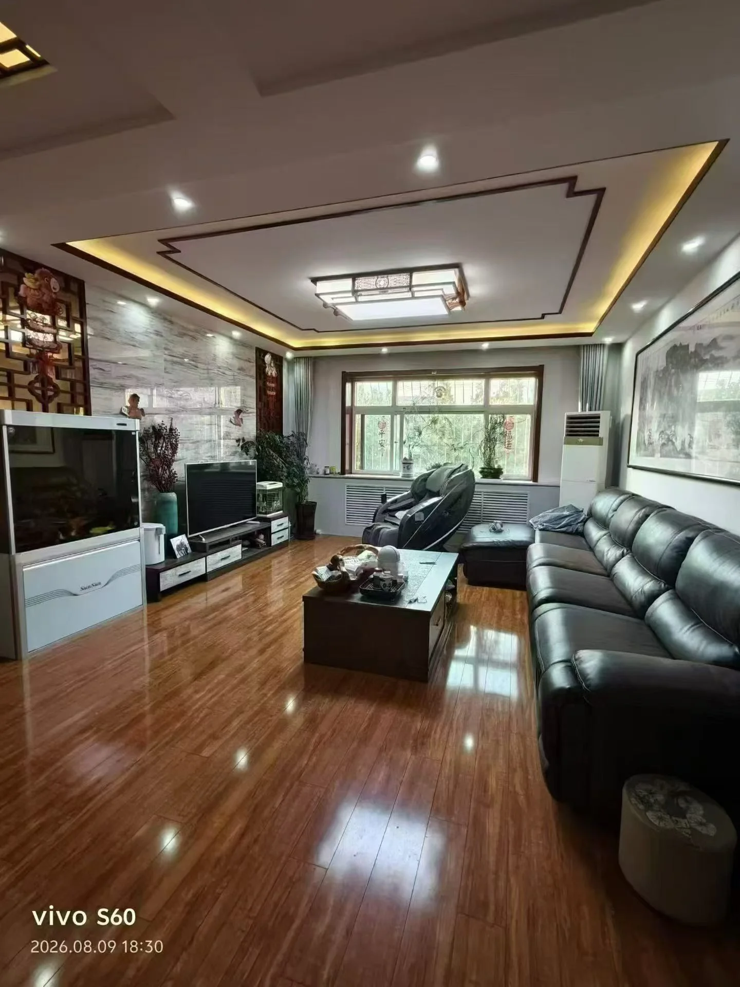 客厅场景，墙面采用现代简约装修风格，配有大型平板电视和深色沙发。采光充足，地面铺设木质地板，整体呈现现代舒适的居住氛围。