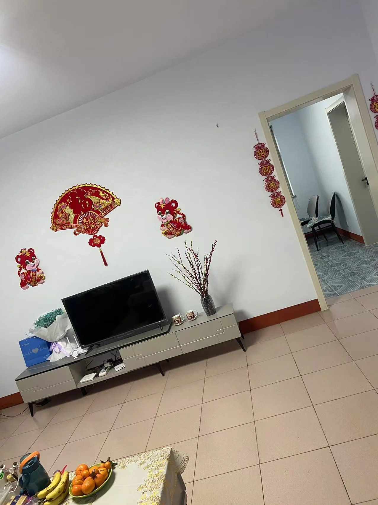 客厅空间，墙面为浅色调，配有电视和简约的电视柜。地面铺设米色瓷砖，整体装修风格偏基础实用型，采光适中。