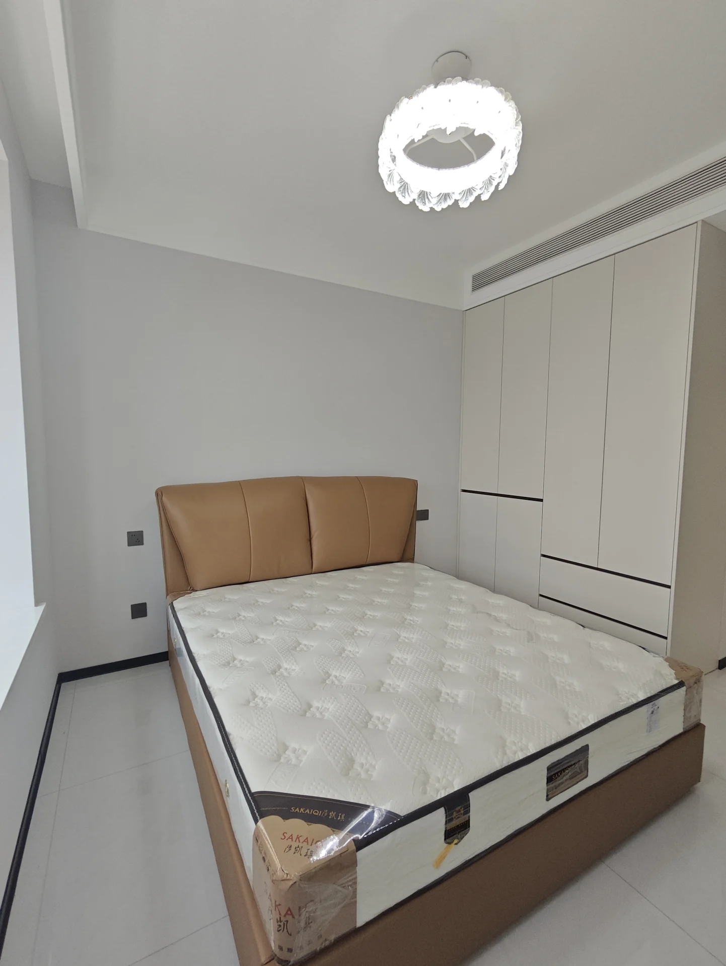 卧室场景，床铺采用米色床品，床头是木质床架，天花板上悬挂着一个带有水晶吊灯的现代吊灯，整体装修风格简洁明亮，光线充足，营造出温馨舒适的卧室氛围。