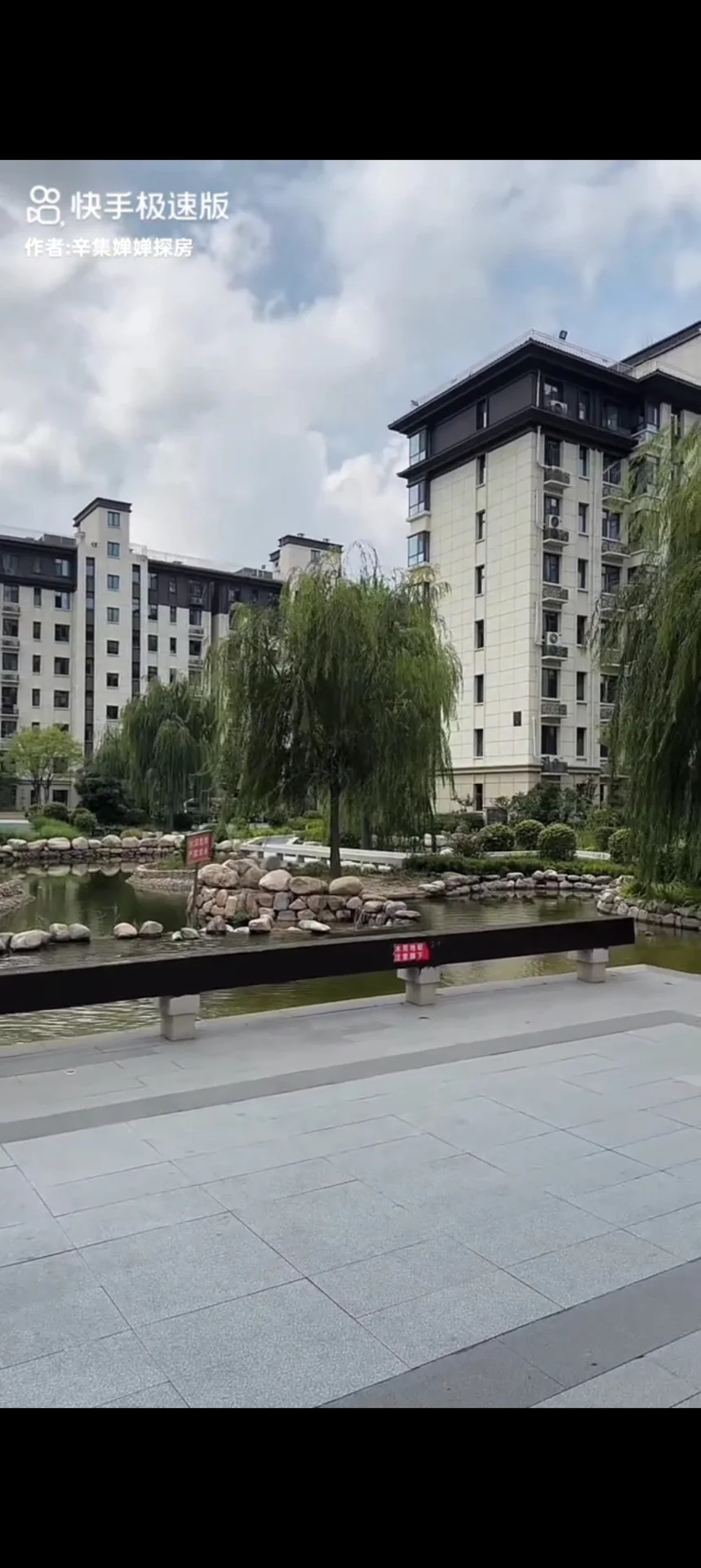 画面展示了多栋现代风格的住宅楼立面，建筑外墙以浅色调为主。前景设有精心布置的水景和茂密的垂柳，整体环境绿化良好，营造出宁静和谐的居住氛围。