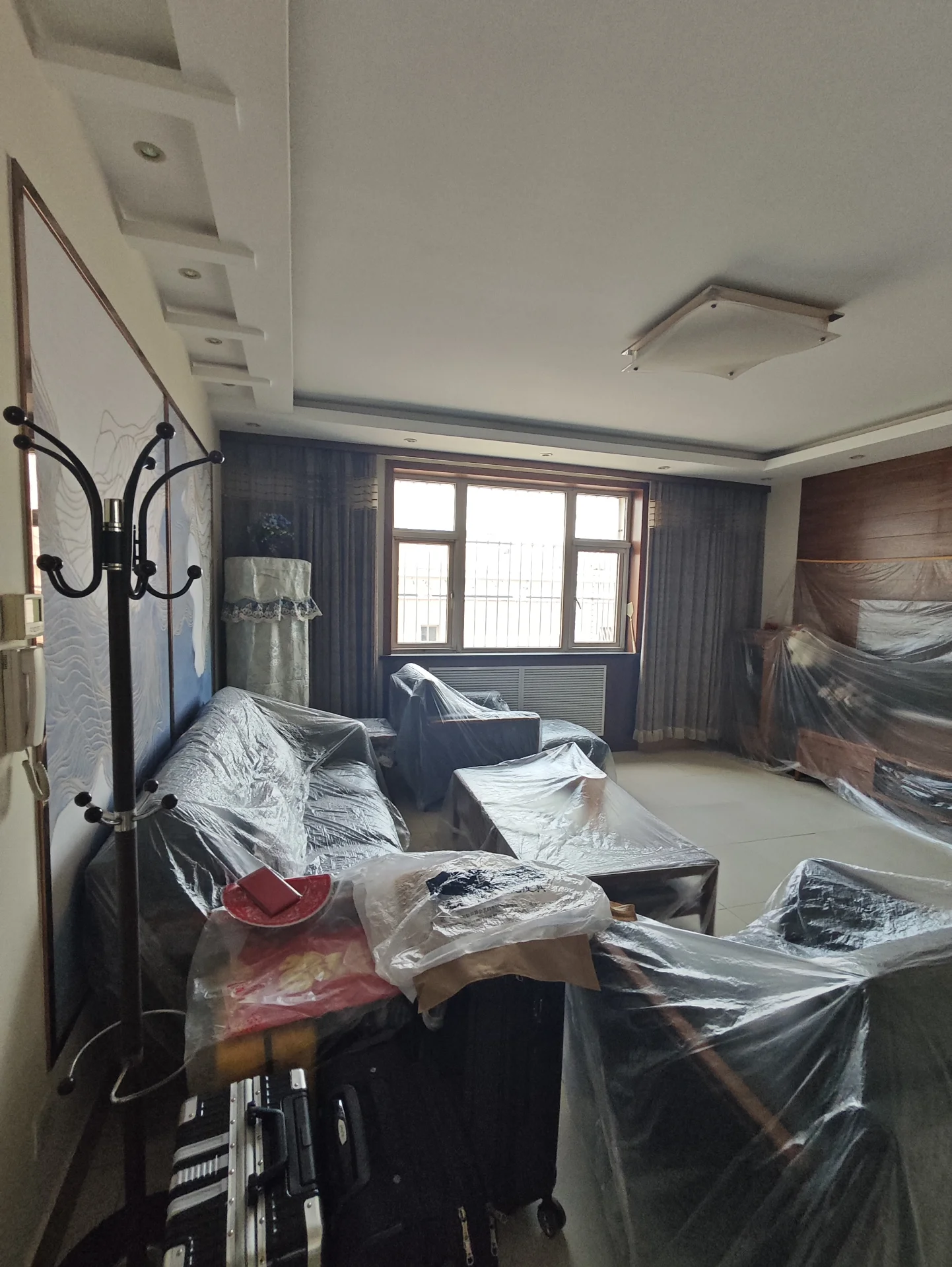 一个室内居住空间，包含多张沙发、床铺以及窗户。空间内有大量的储物箱和杂物堆放，光线透过窗户进入，整体呈现一种生活化、略显凌乱的居住氛围。