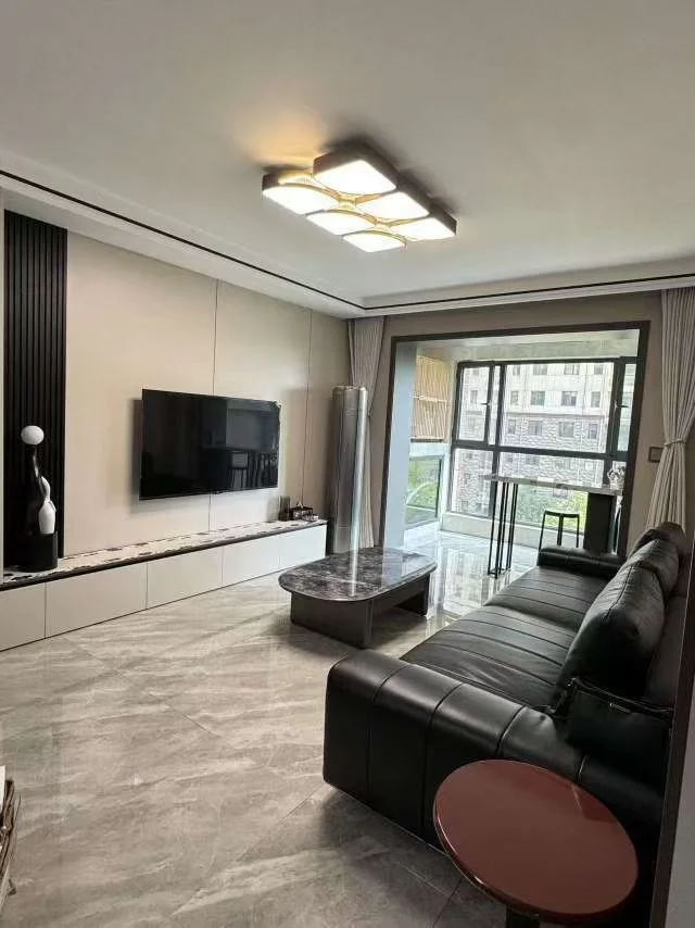 客厅场景，墙面采用灰白色调，配有大型平板电视和简约沙发。采光充足，整体呈现现代简约装修风格，氛围整洁舒适。