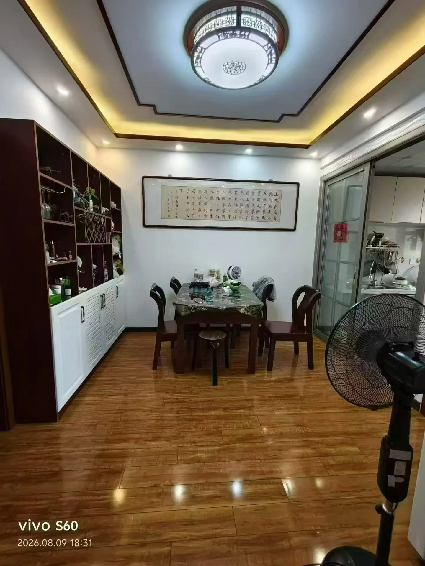 一个室内环境图，以木质地板和木质书架为主要元素，配有木质家具和吊顶。整体装修风格偏向传统或温馨居家风格，光线柔和，营造出一种温馨、略显陈旧的居家氛围。