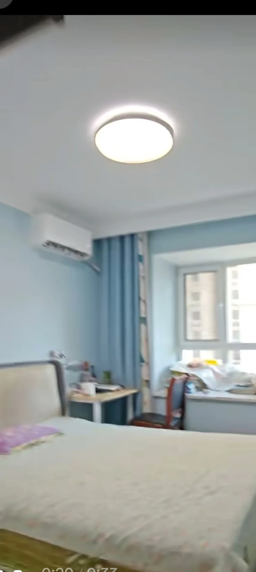 卧室场景，墙面采用柔和的蓝色调，配有床铺和吊扇等基础家具。采光良好，整体呈现简洁、清爽的居住氛围。
