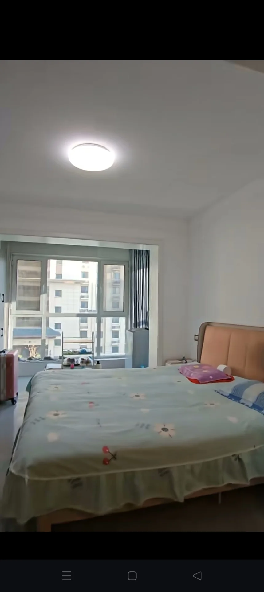 卧室场景，床铺铺着浅色花纹床单，房间采光良好，窗户提供了充足的自然光，整体呈现温馨、整洁的卧室居住氛围。