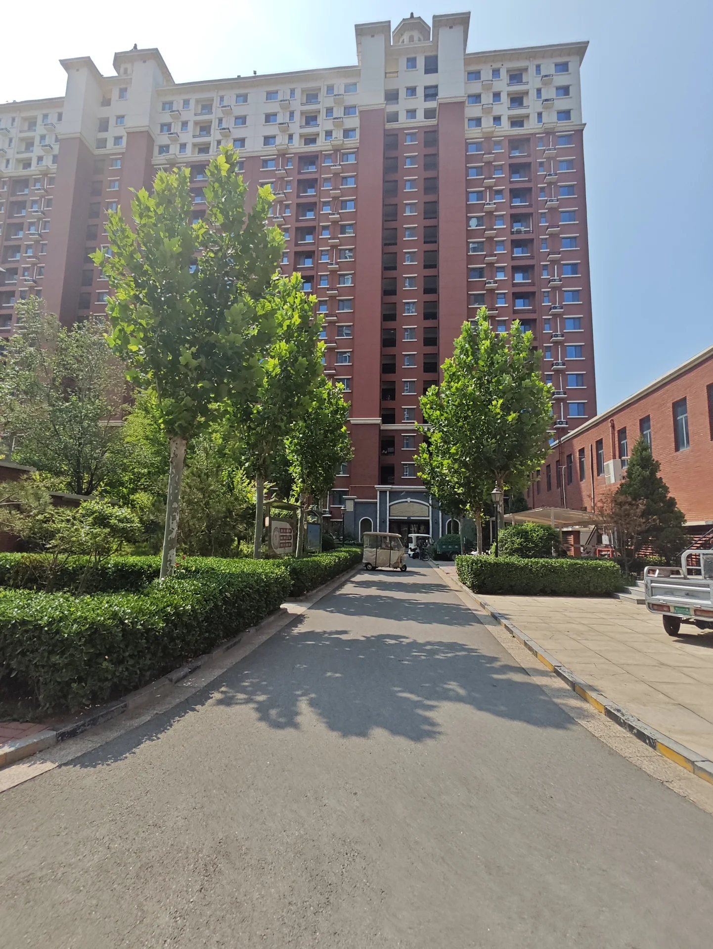 一栋多层住宅楼的外部立面，建筑主体呈现红棕色调，设有多个阳台和窗户。建筑前方有整齐的绿化带和道路，整体呈现出社区住宅的宁静和谐氛围。