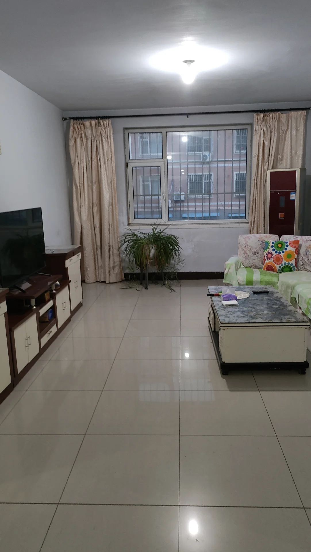 客厅场景，地面铺设米色瓷砖，配有大型平板电视和简约沙发。采光良好，整体呈现现代简约装修风格，氛围整洁舒适。