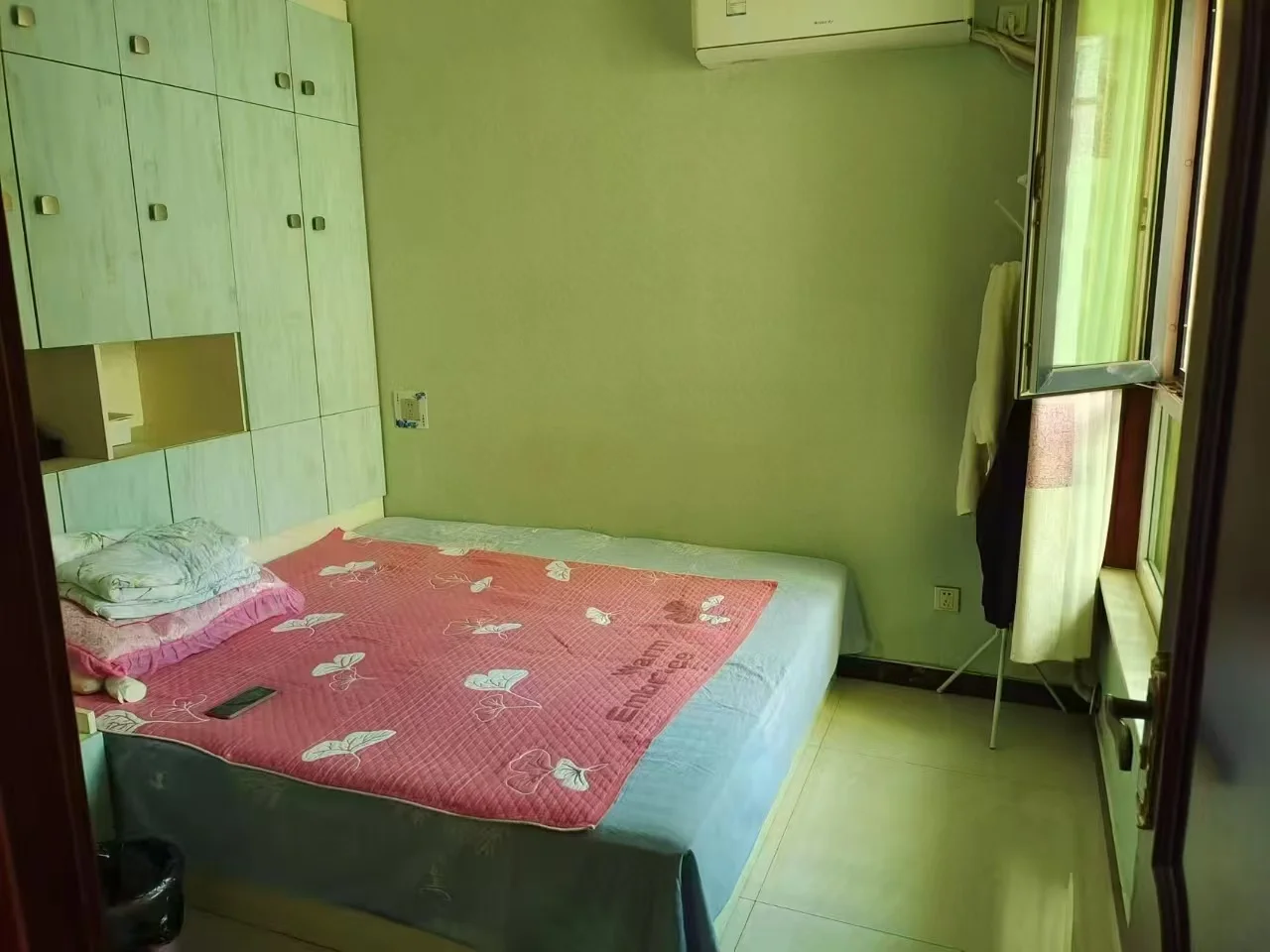 卧室场景，墙面为浅绿色，配有木质衣柜和床铺。采光良好，整体呈现简洁、温馨的居住氛围。